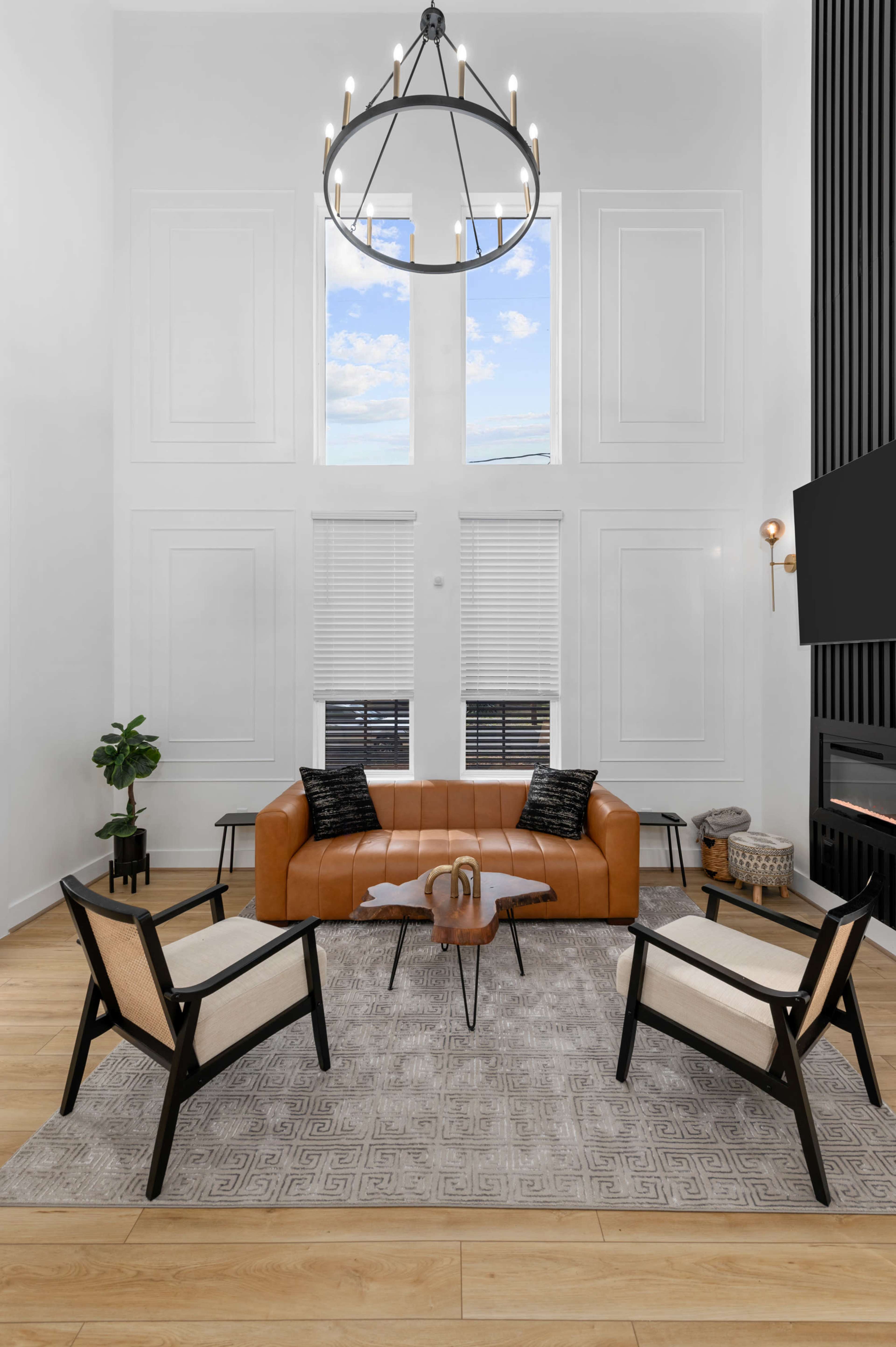 The living room features a high ceiling, large windows, a brown leather sofa, and two black and beige chairs surrounding a central glass coffee table.