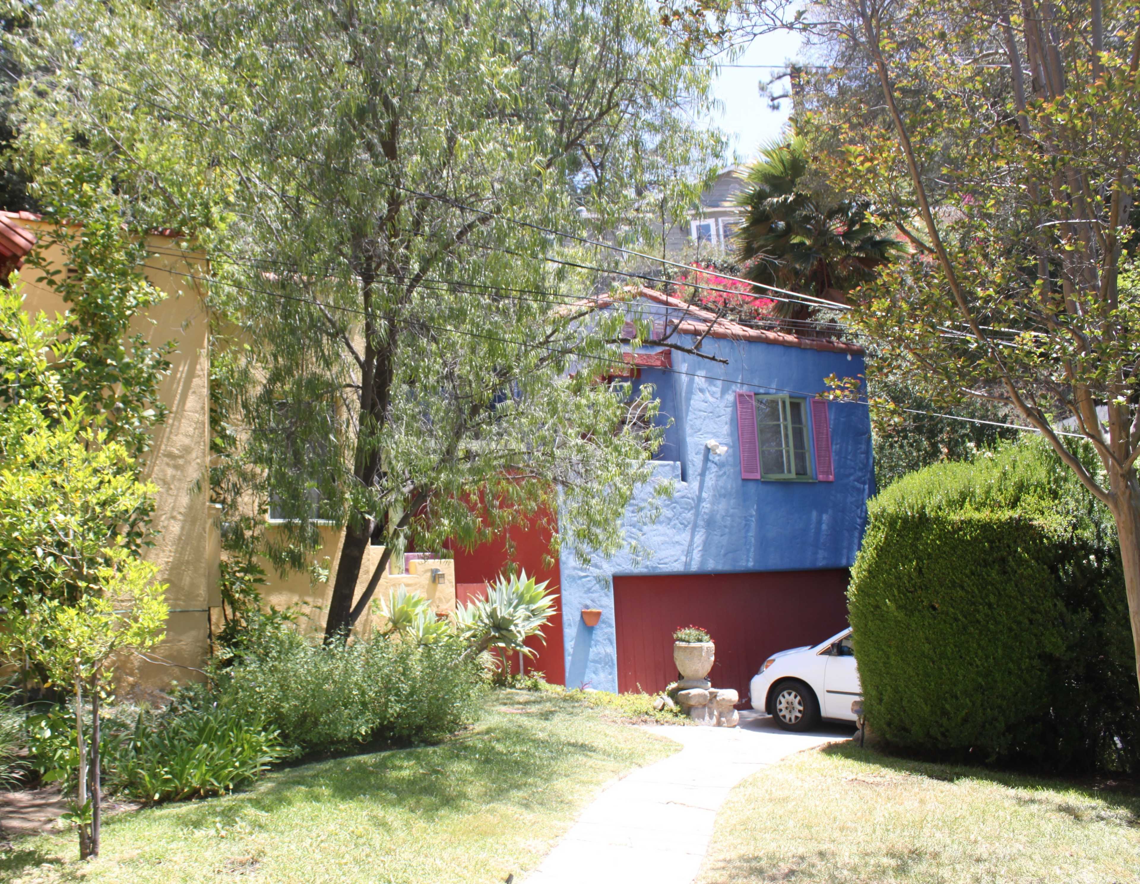 The image shows a winding path leading past two colorful houses, with greenery and shrubs lining the way.