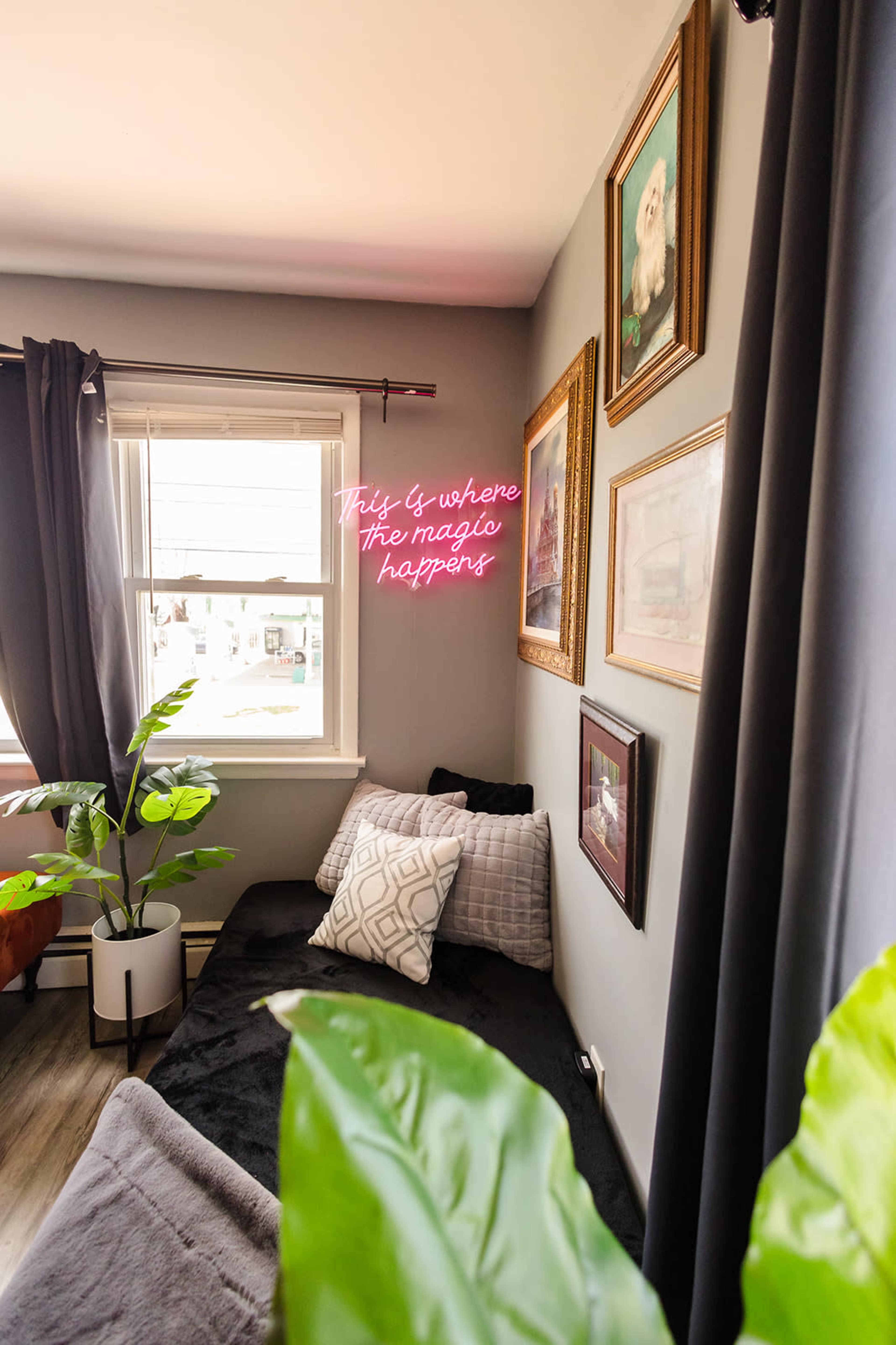 A cozy room featuring a dark couch, decorative framed artwork on the walls, and a neon sign that reads "This is where the magic happens" near a window.