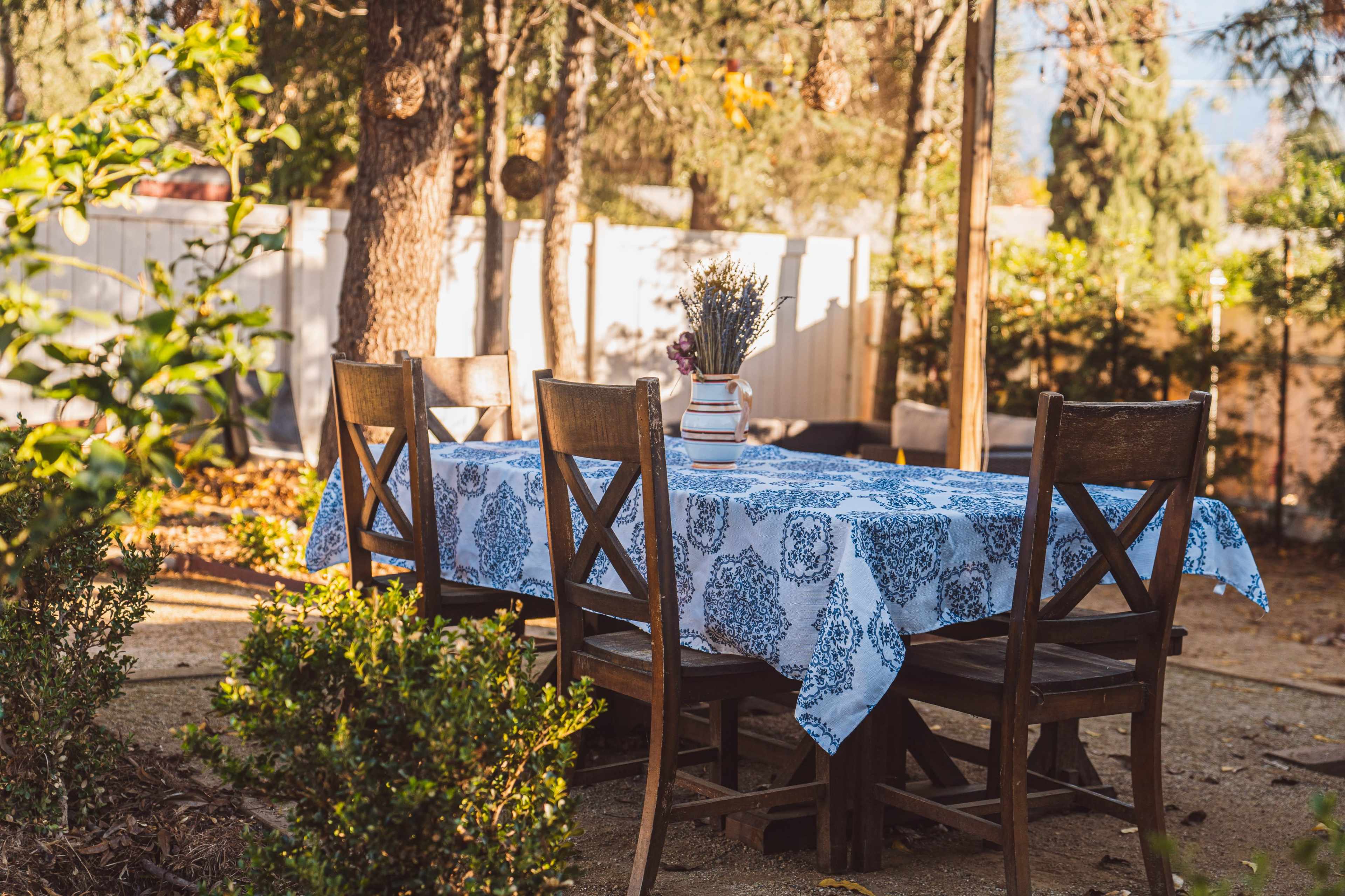 A table with a blue patterned tablecloth is set under trees in a garden, surrounded by wooden chairs and greenery.