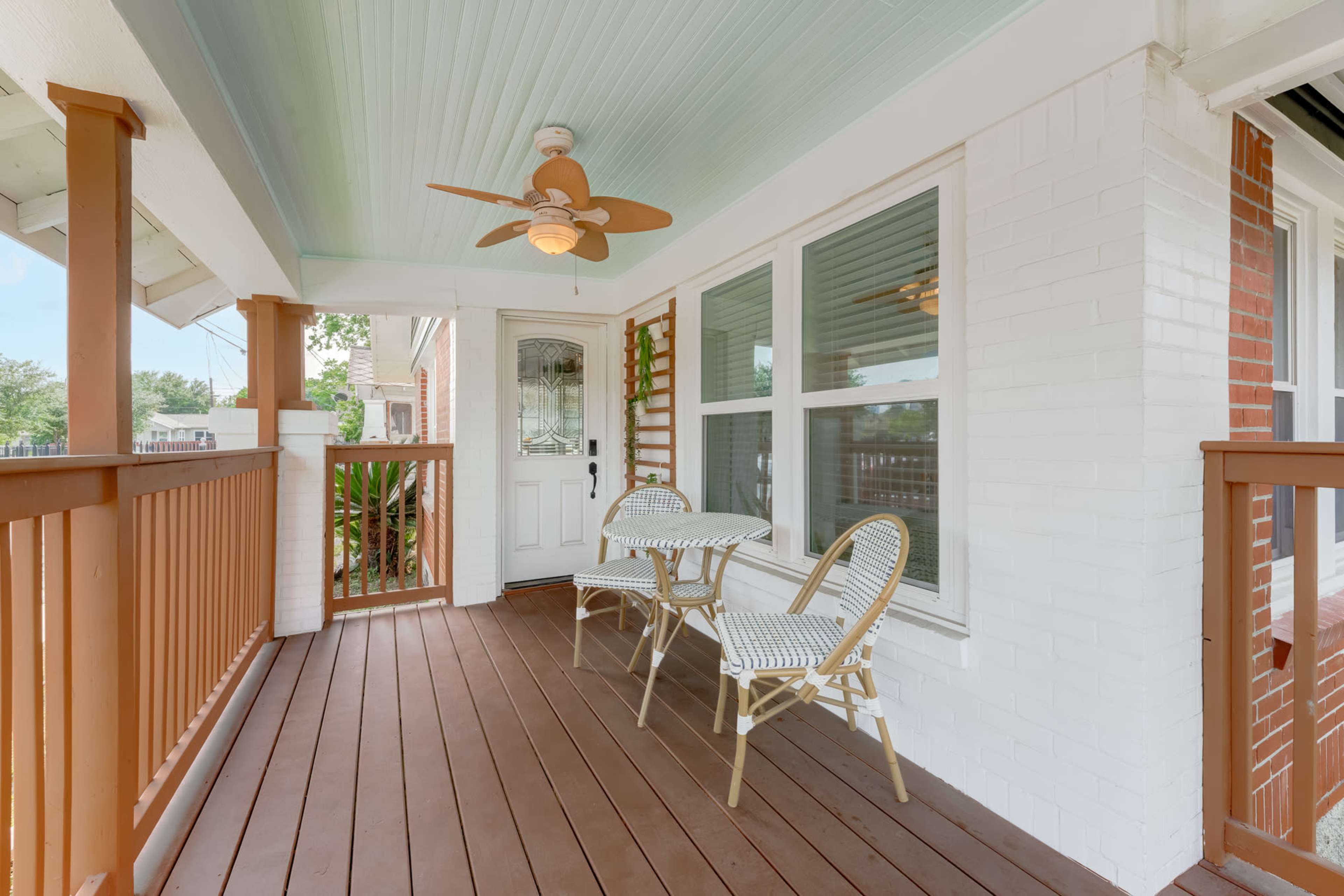A covered porch with two chairs and a table, featuring a ceiling fan and light fixture, surrounded by large windows.