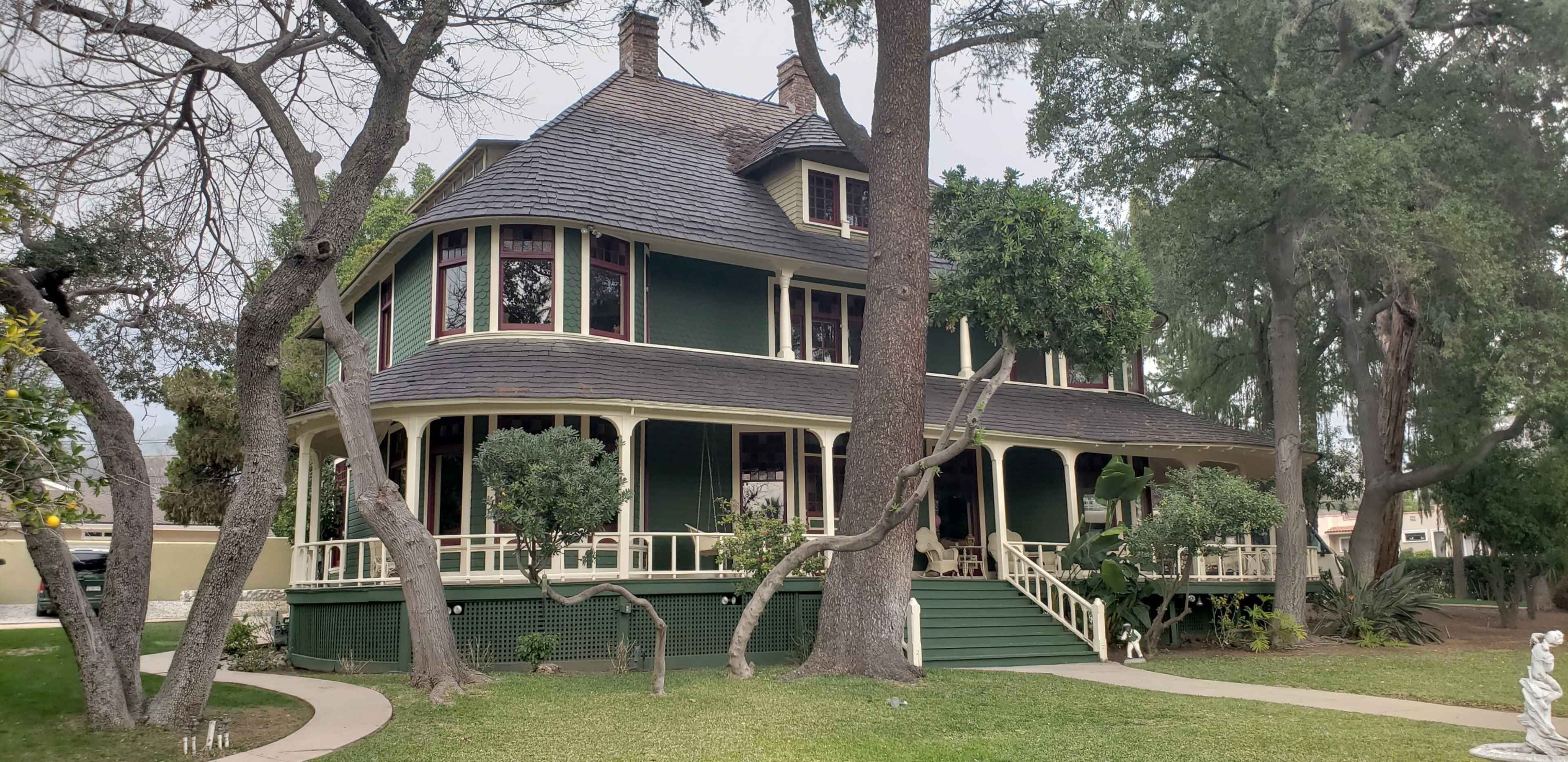 A large green Victorian-style house with a wraparound porch, surrounded by trees and landscaped grass.