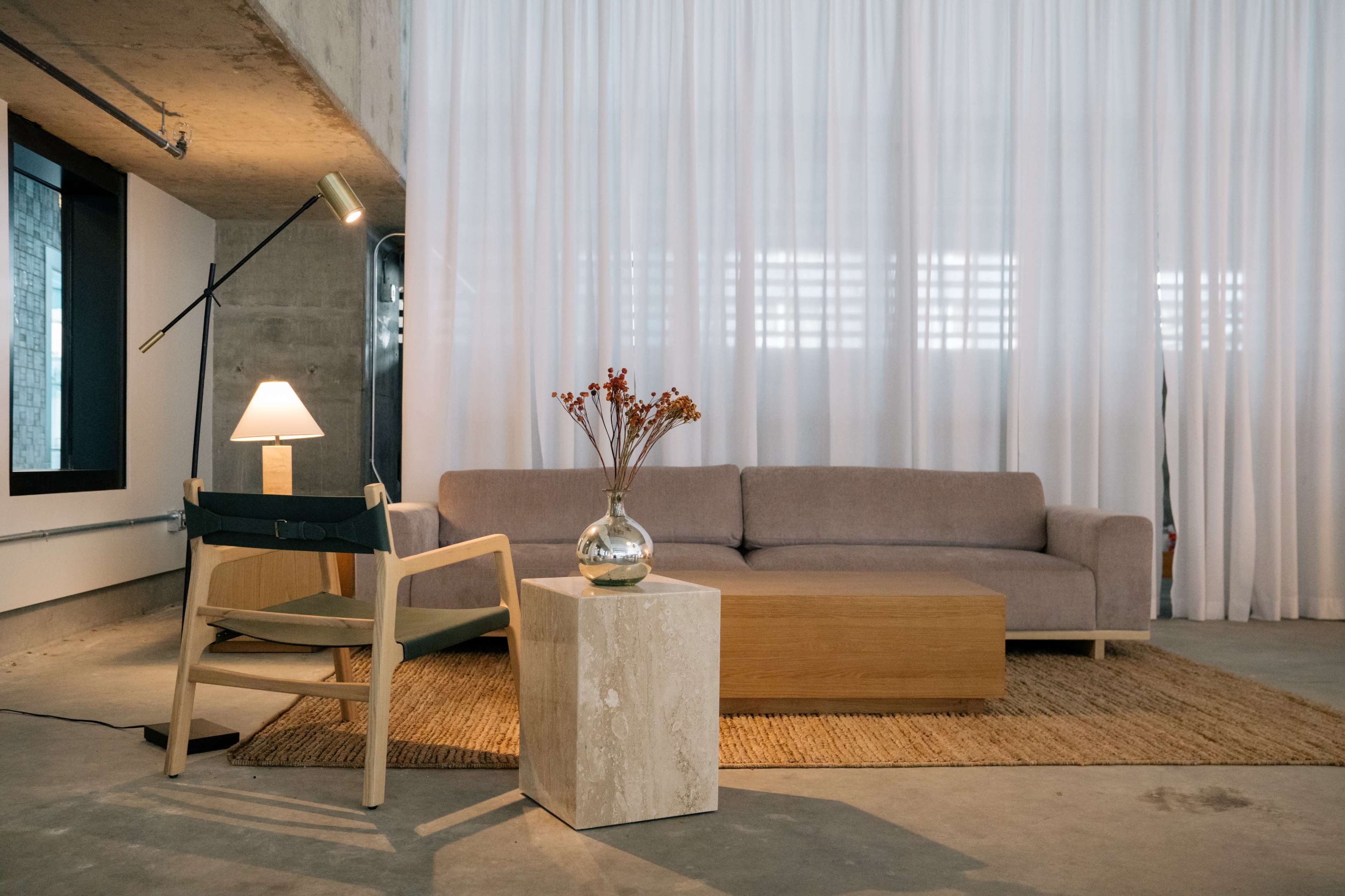The image shows a modern living area with a sofa, a wooden coffee table, a chair, a lamp, and a vase of dried flowers against a backdrop of sheer curtains.