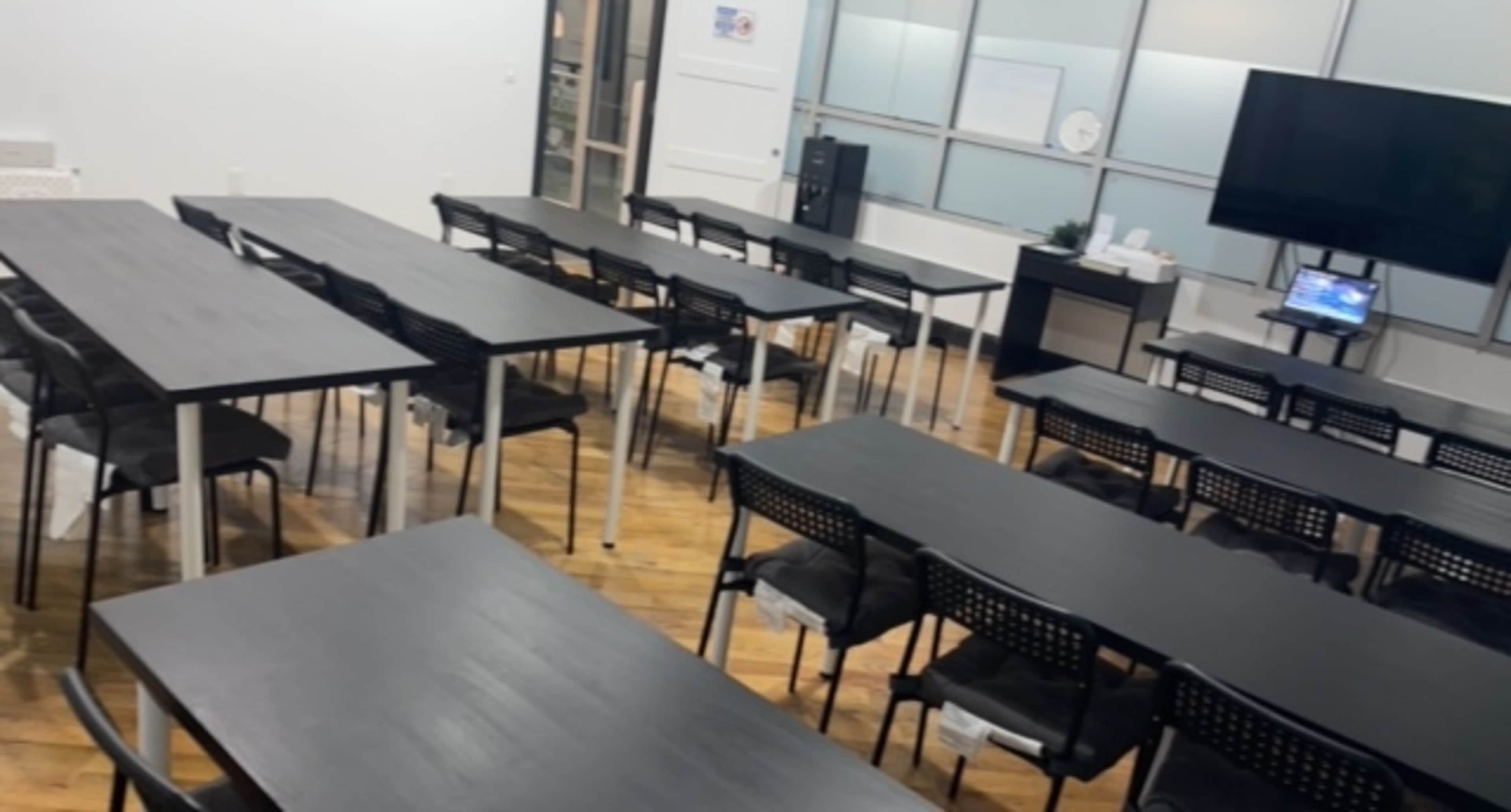 A classroom or meeting space arranged with multiple black tables and chairs, with a television and a computer set up at the front.