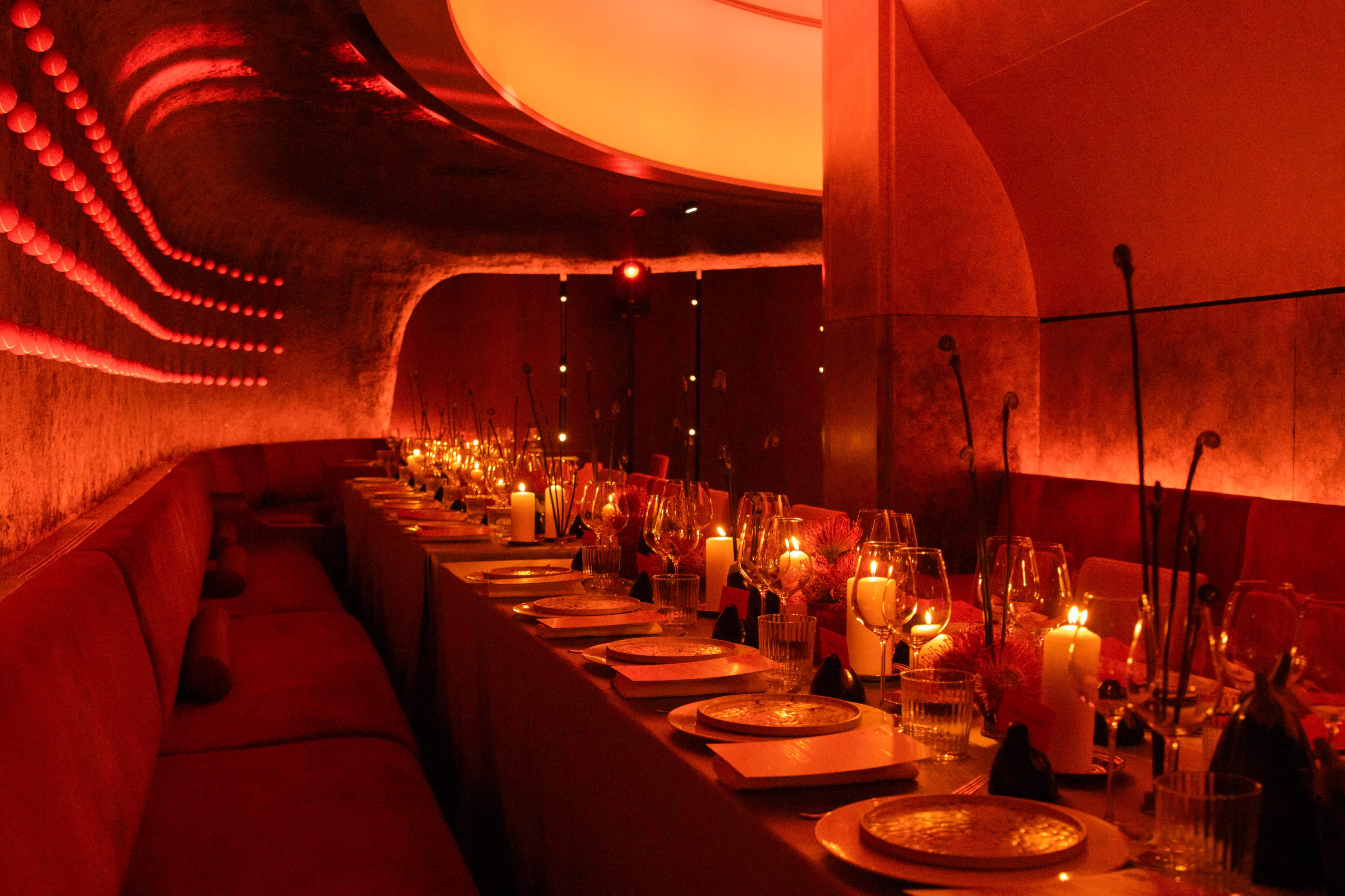 The image shows a dimly lit restaurant interior featuring a long dining table set with elegant tableware and numerous candles.
