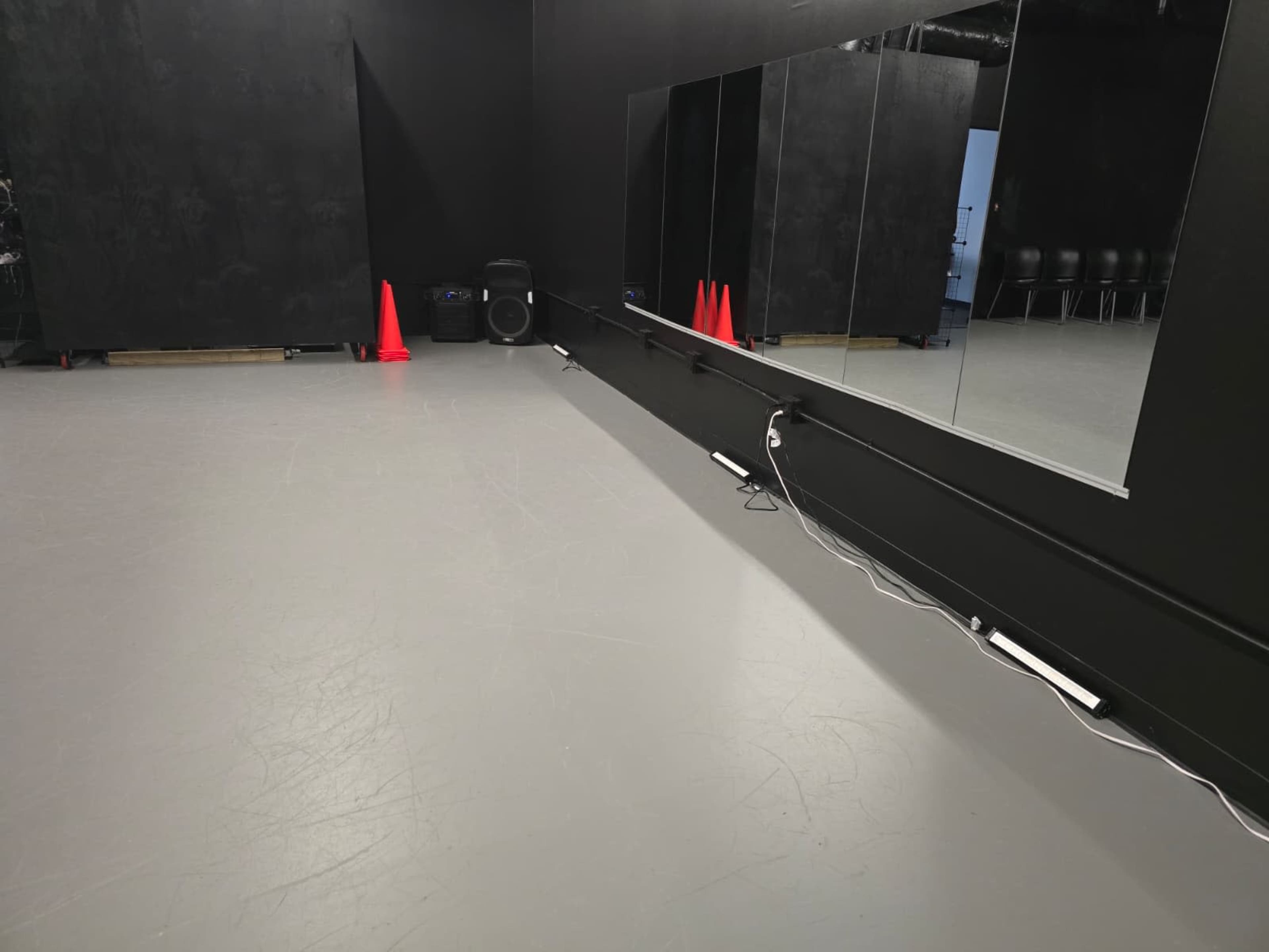 The image shows a dimly lit dance studio with a gray floor, a large mirror on one wall, and red cones placed on the floor.