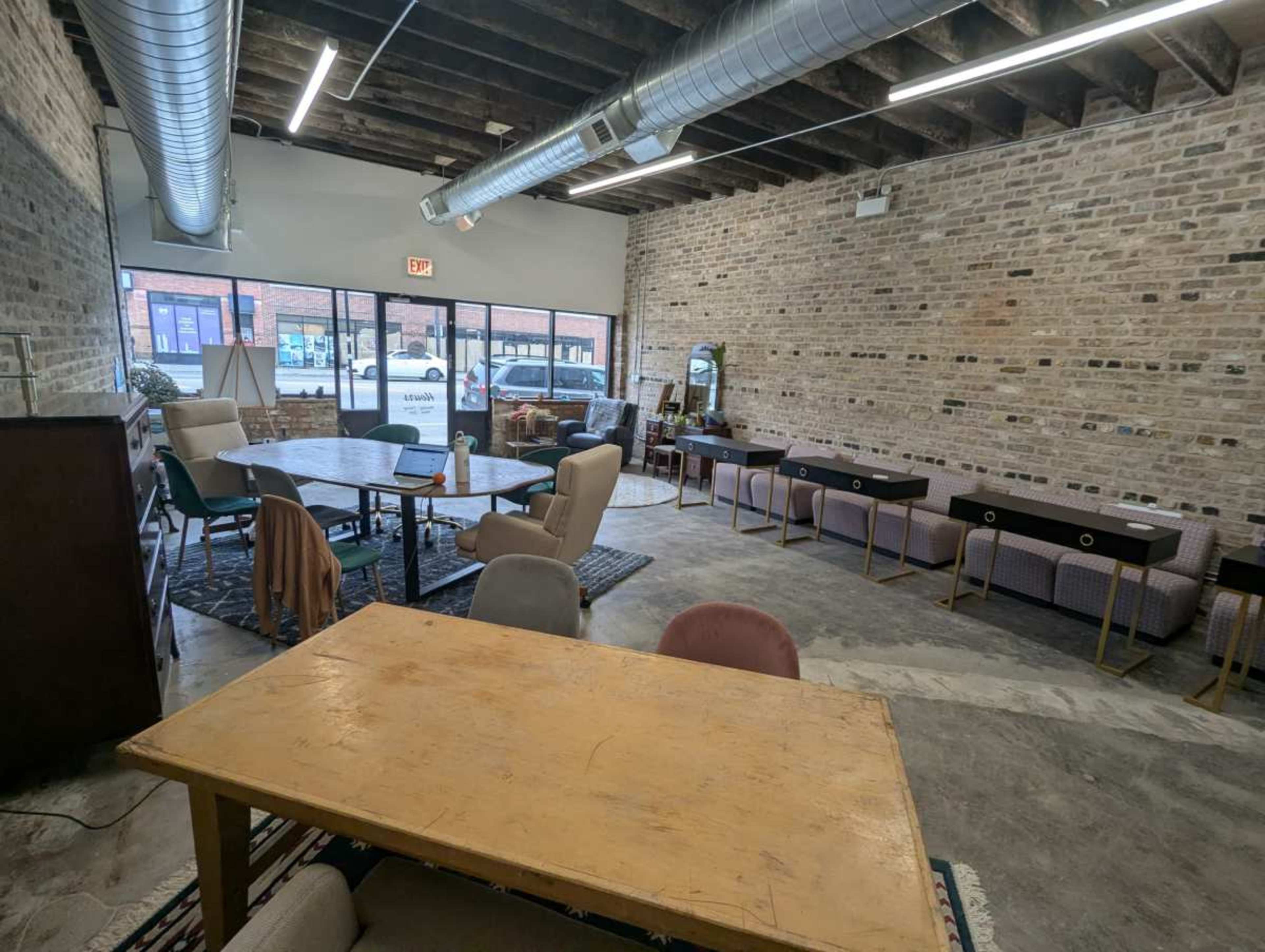 The image shows a spacious, minimally furnished room with exposed brick walls, wooden beams, and a mix of tables and chairs arranged throughout the area.