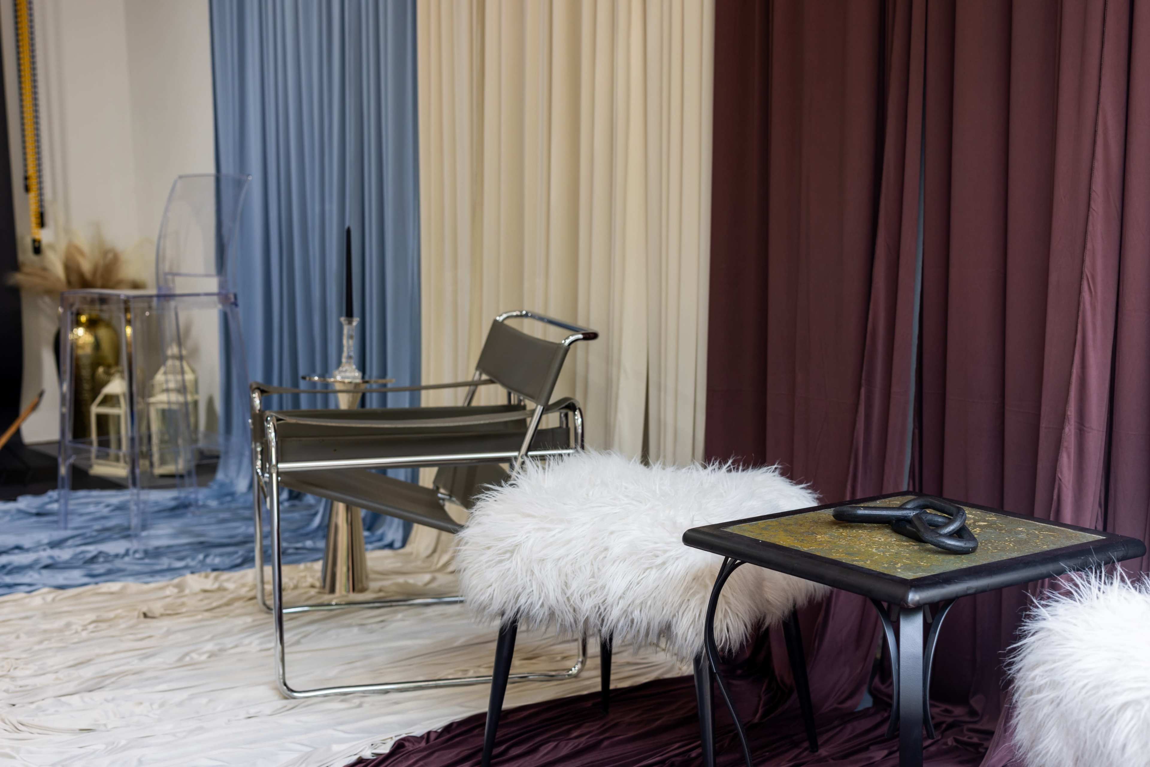 The image shows a cozy interior setting with floor drapery in varying colors, a modern chair with a fur seat, and a small table with a decorative object.