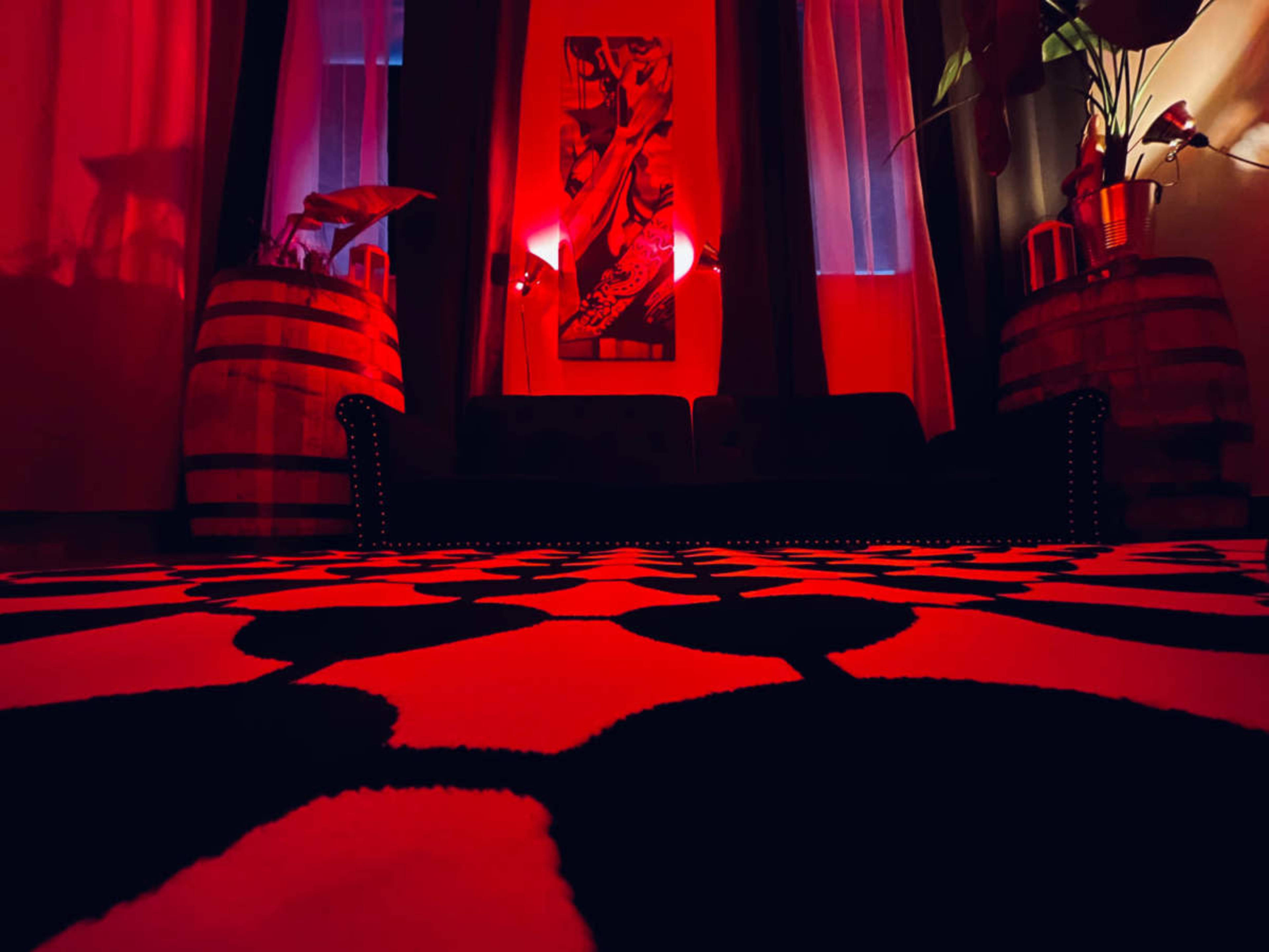 The image shows a room with a patterned black and white rug, a dark couch, and two wooden barrels under dim red lighting.
