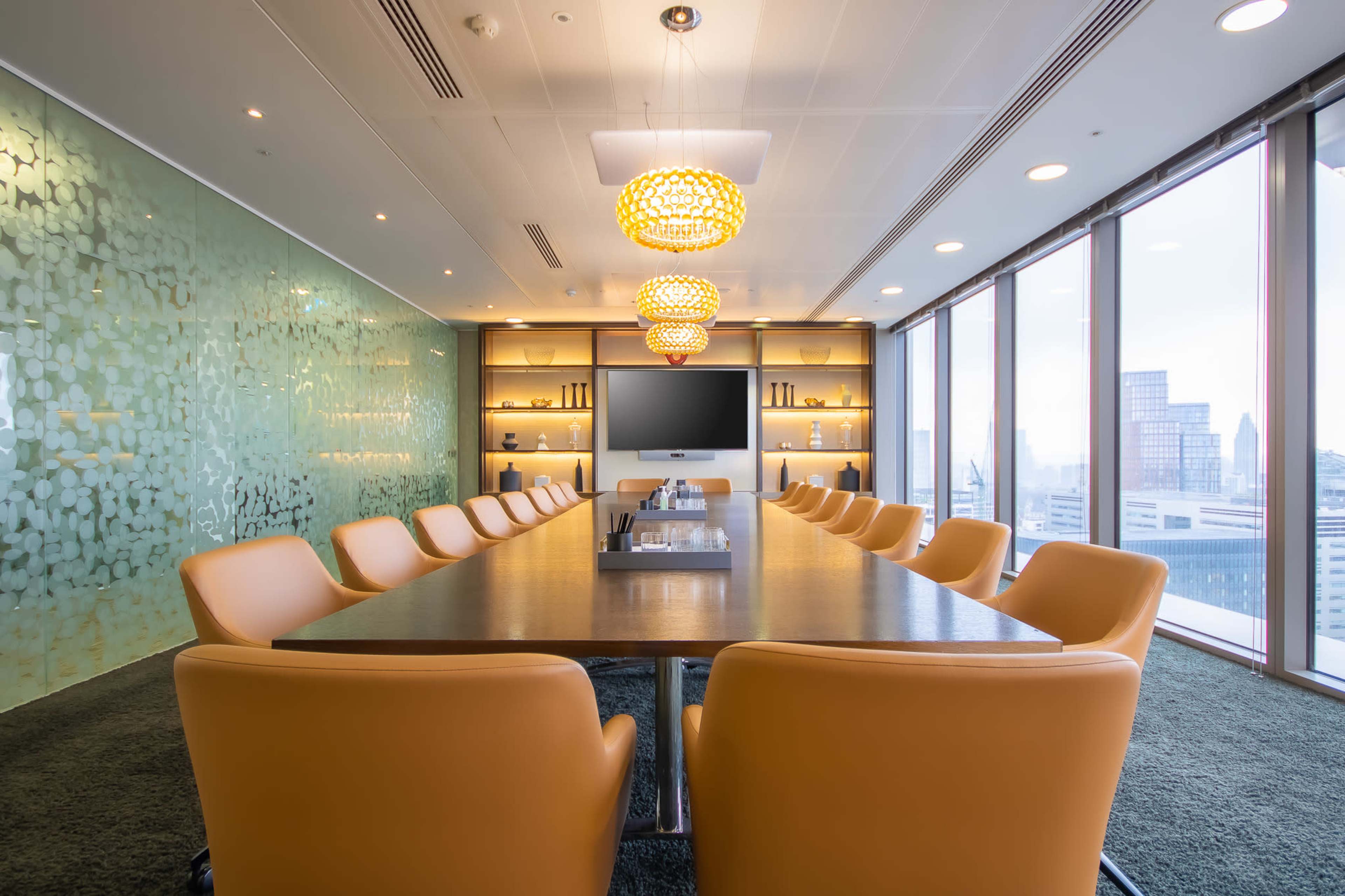 A modern conference room features a long wooden table surrounded by orange chairs, with large windows offering a view of the city and decorative wall elements.