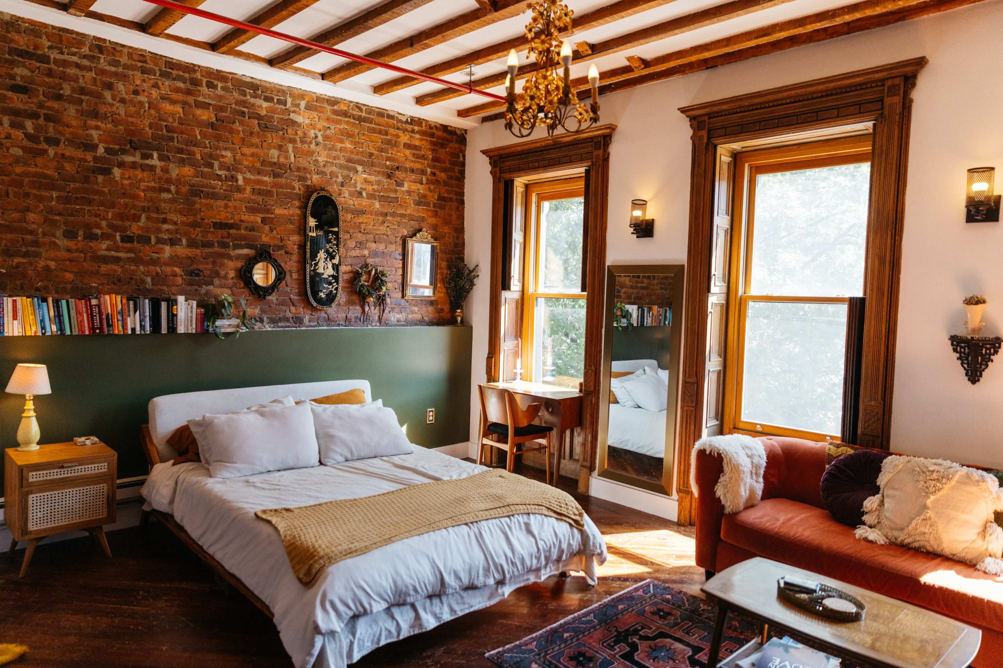A well-lit bedroom features a mix of exposed brick walls, a bed with white linens, a wooden table, and a cozy seating area.