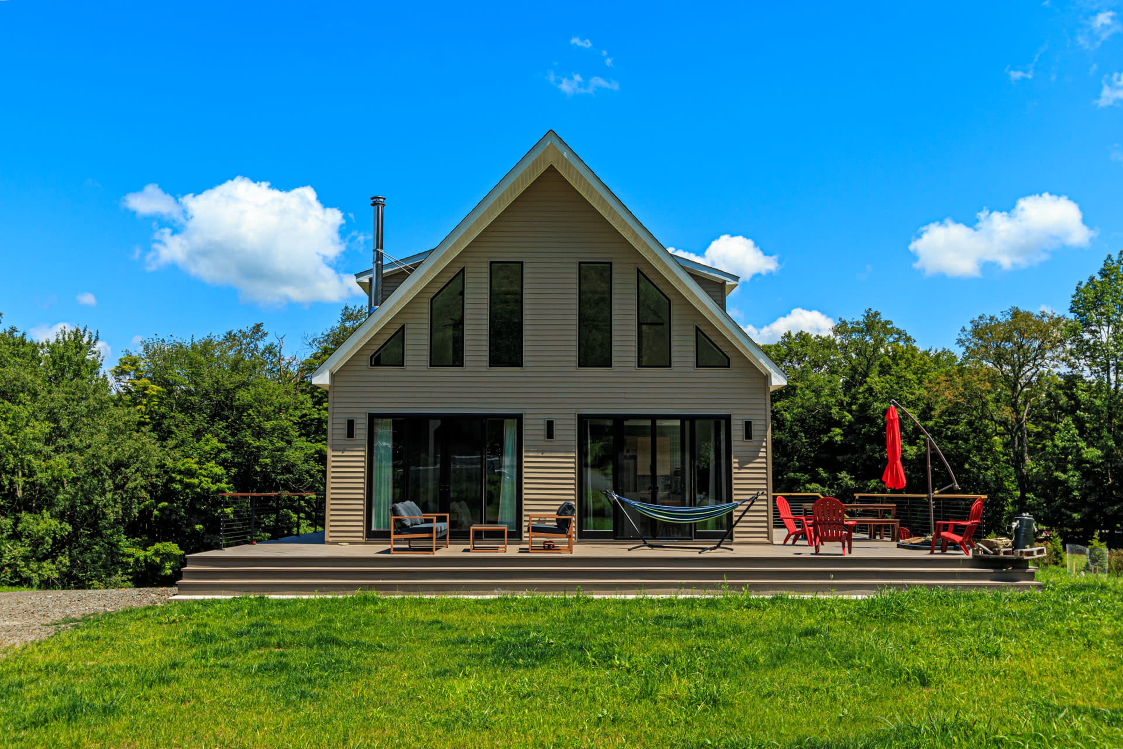 The image shows a modern house with large windows and a deck featuring red outdoor chairs, a hammock, and a red umbrella, set against a blue sky and greenery.
