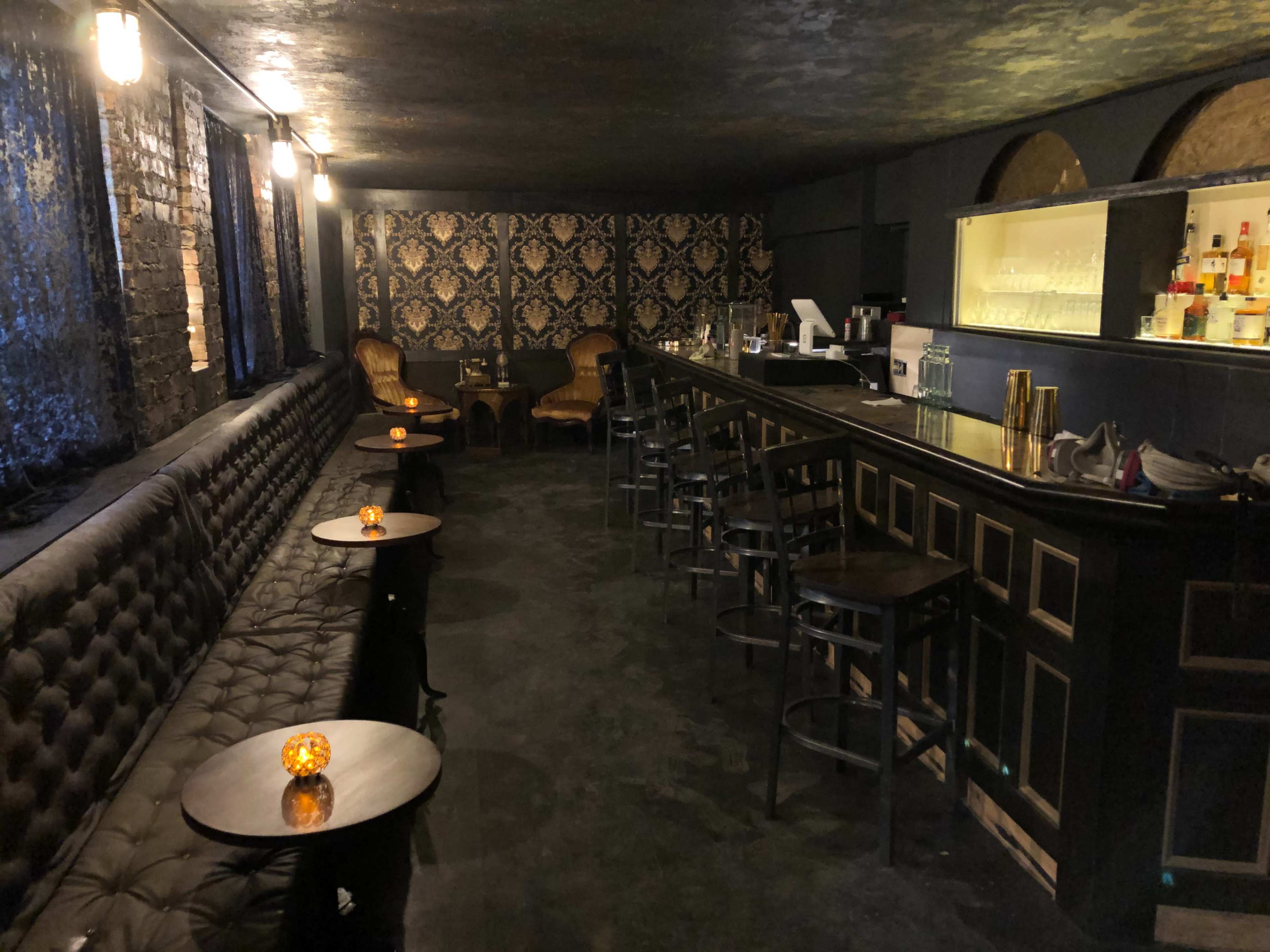 A dimly lit bar with tufted seating, small tables, and a counter stocked with drinks.