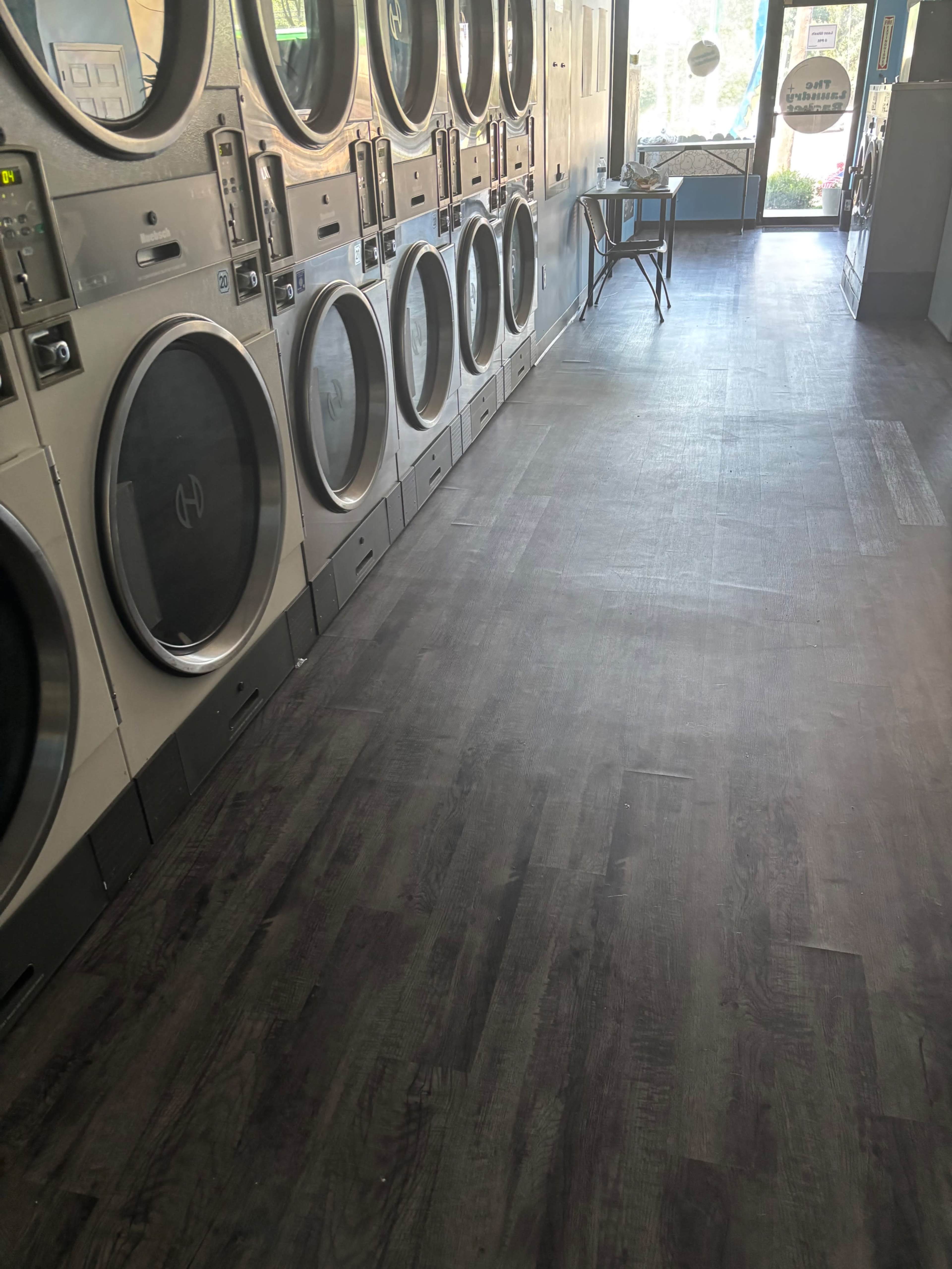 The image shows the interior of a laundromat with multiple washing machines aligned against the wall and a folding table in the middle of the room.