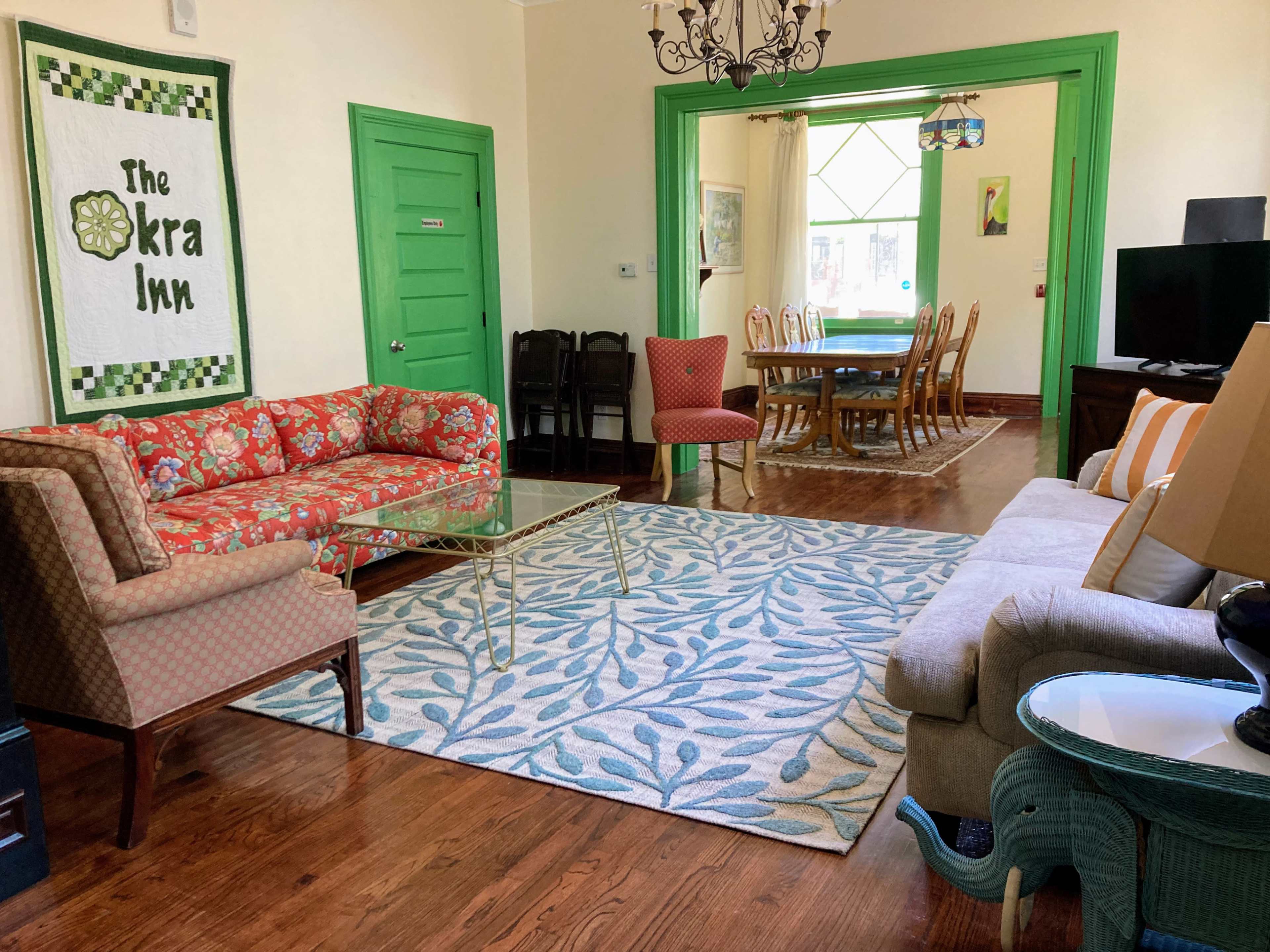 The image shows a brightly colored living room with two patterned sofas, a glass coffee table, a decorative rug, and a dining area visible through an open doorway.