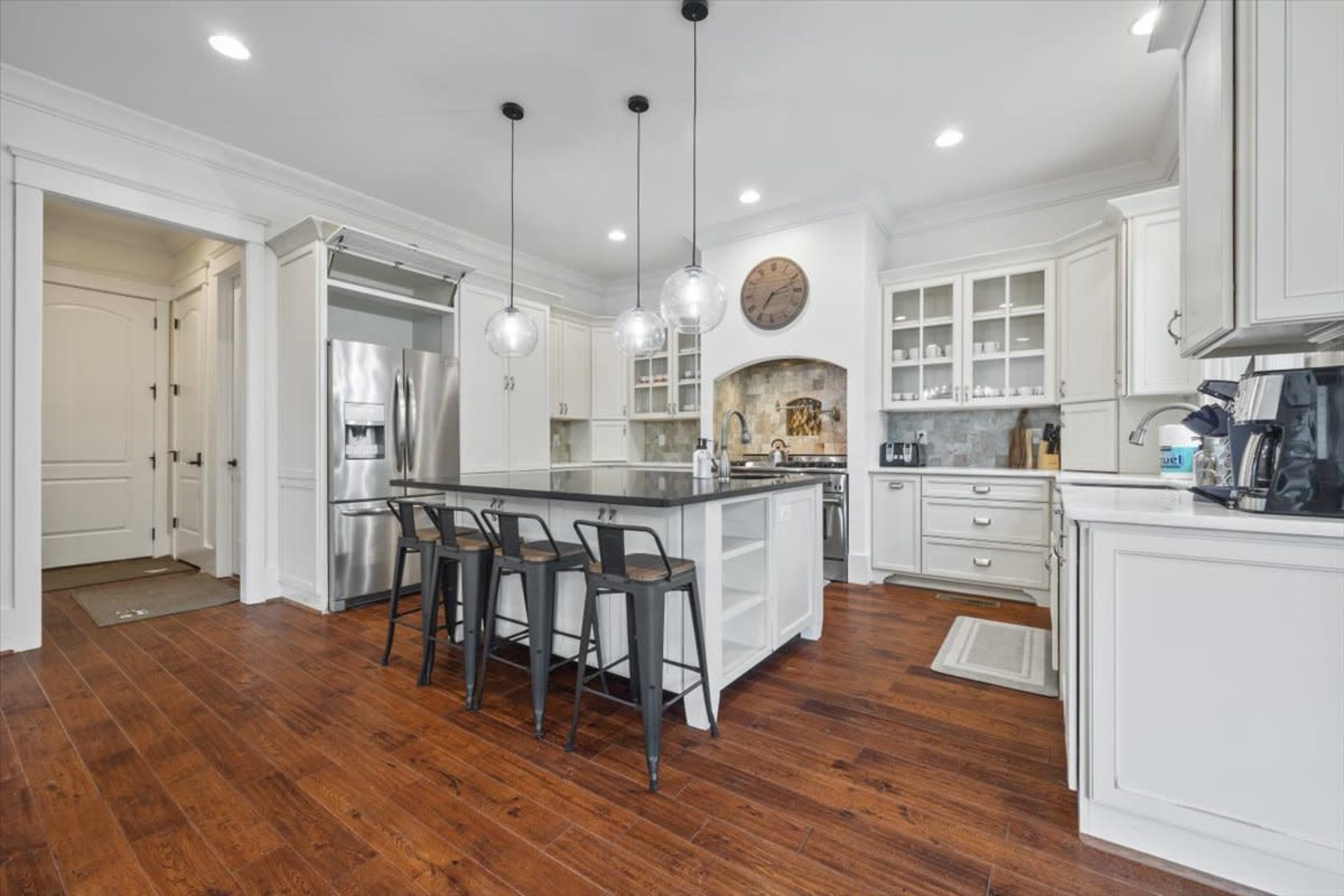 A modern kitchen features a central island with three stools, white cabinetry, and hardwood flooring.