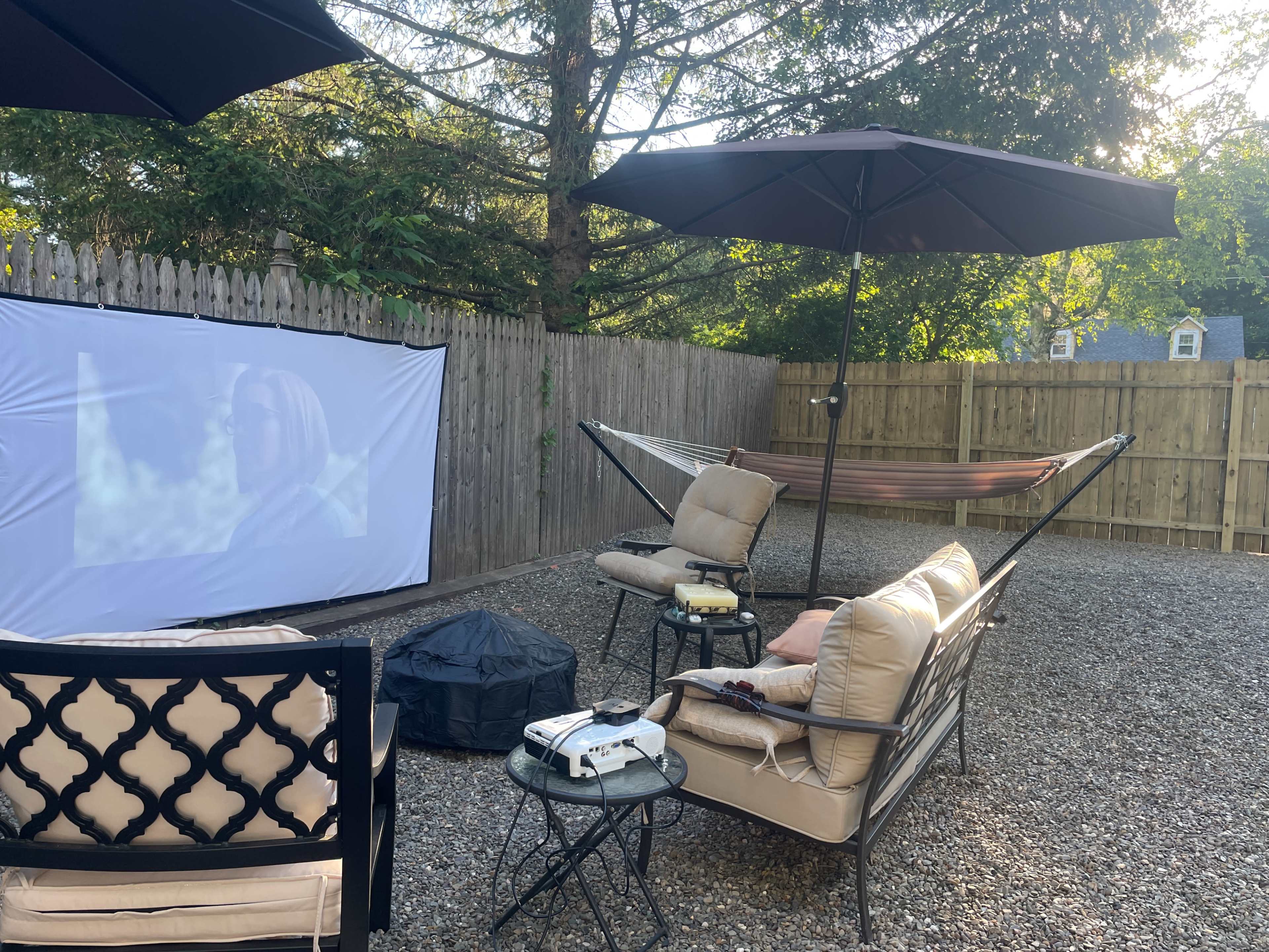 The image shows a backyard patio area with a portable projector screen, seating arrangements, a hammock, and two black umbrellas.