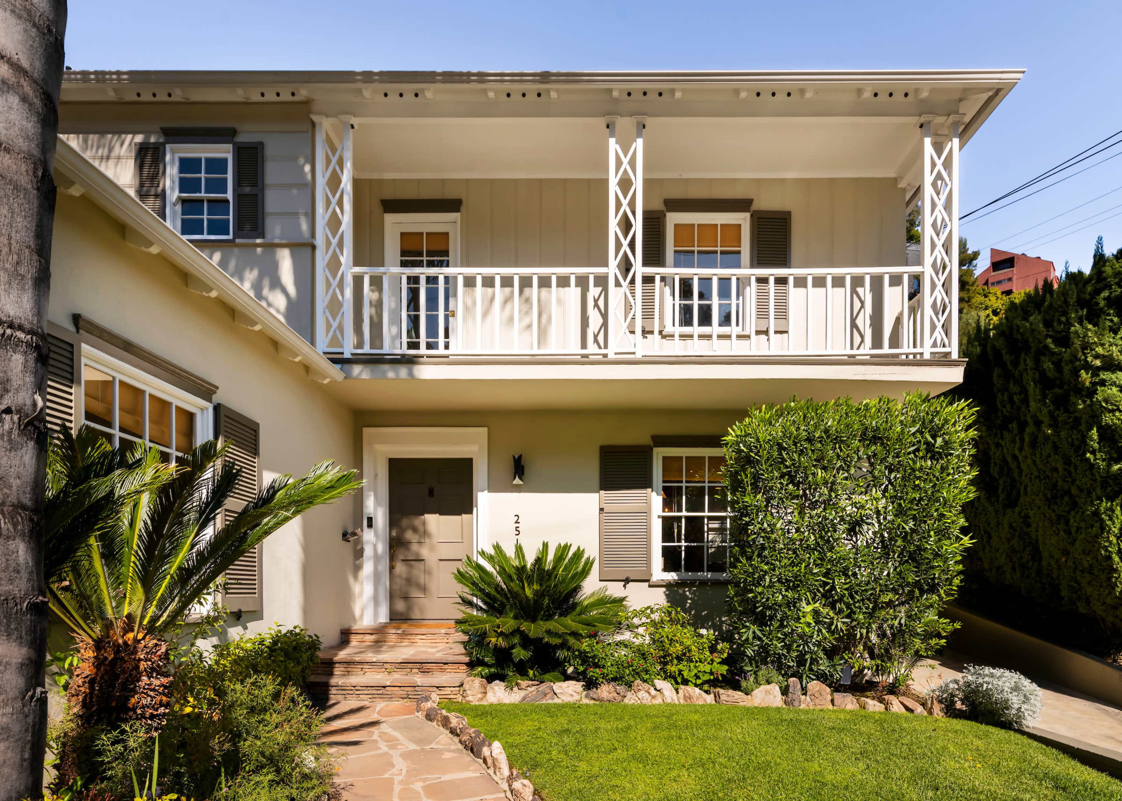 The image features a two-story house with a front porch, surrounded by lush greenery and a stone pathway leading to the entrance.