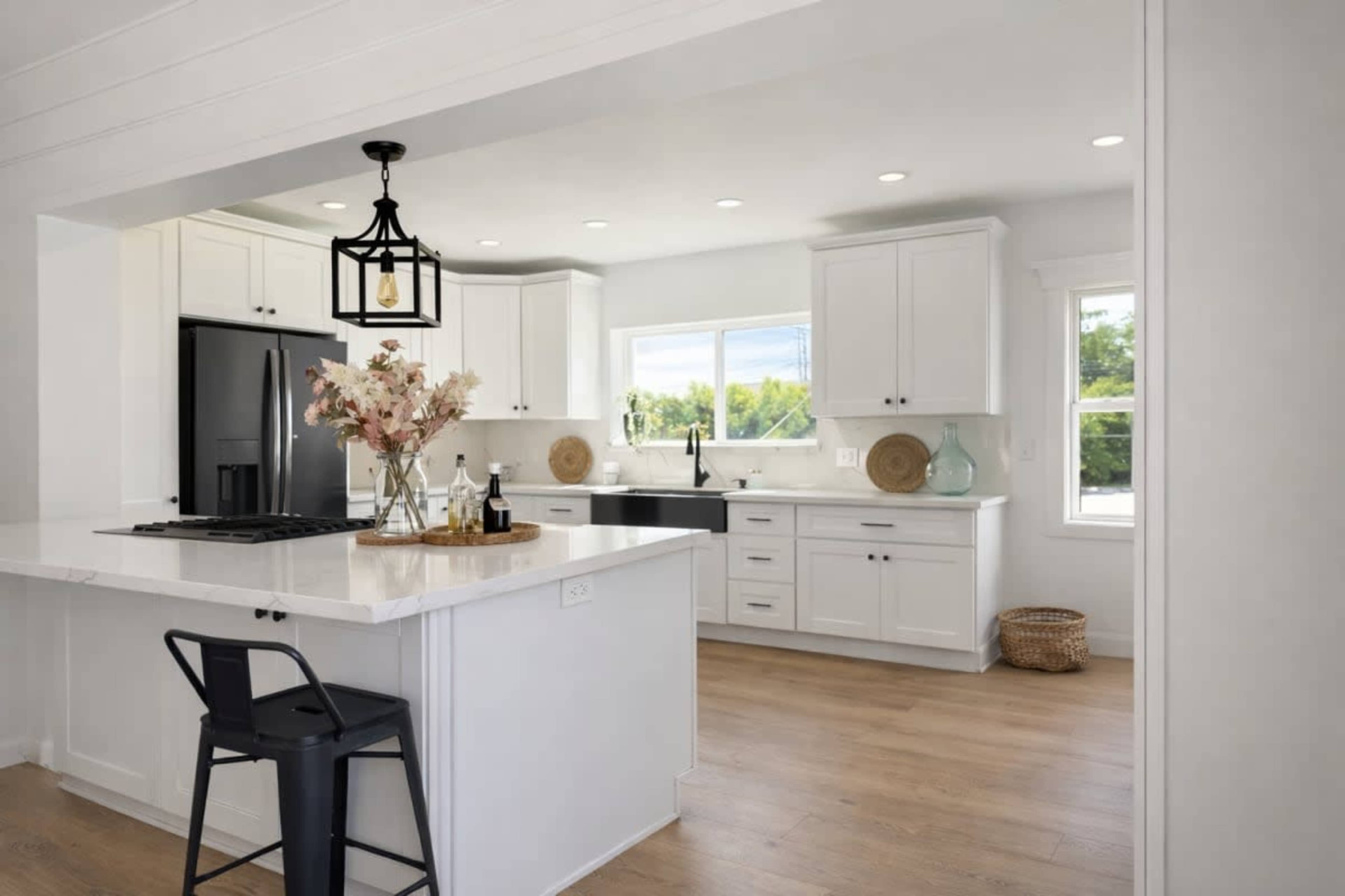 The image shows a bright modern kitchen featuring white cabinetry, a central island with seating, and large windows that allow natural light to fill the space.