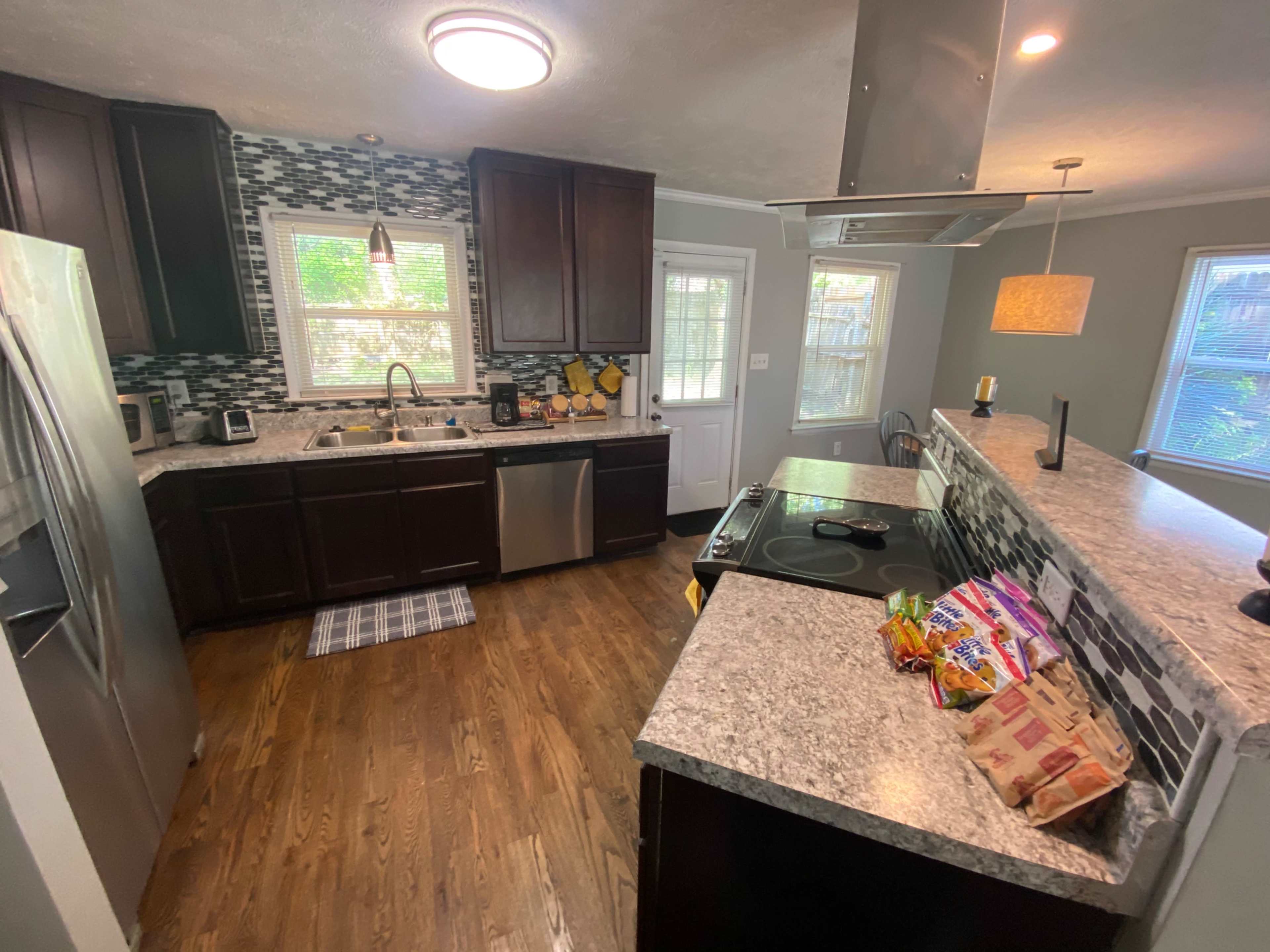 A modern kitchen featuring dark cabinets, a stainless steel refrigerator, and a granite countertop with a small breakfast bar, along with a view of a backyard through a door and window.