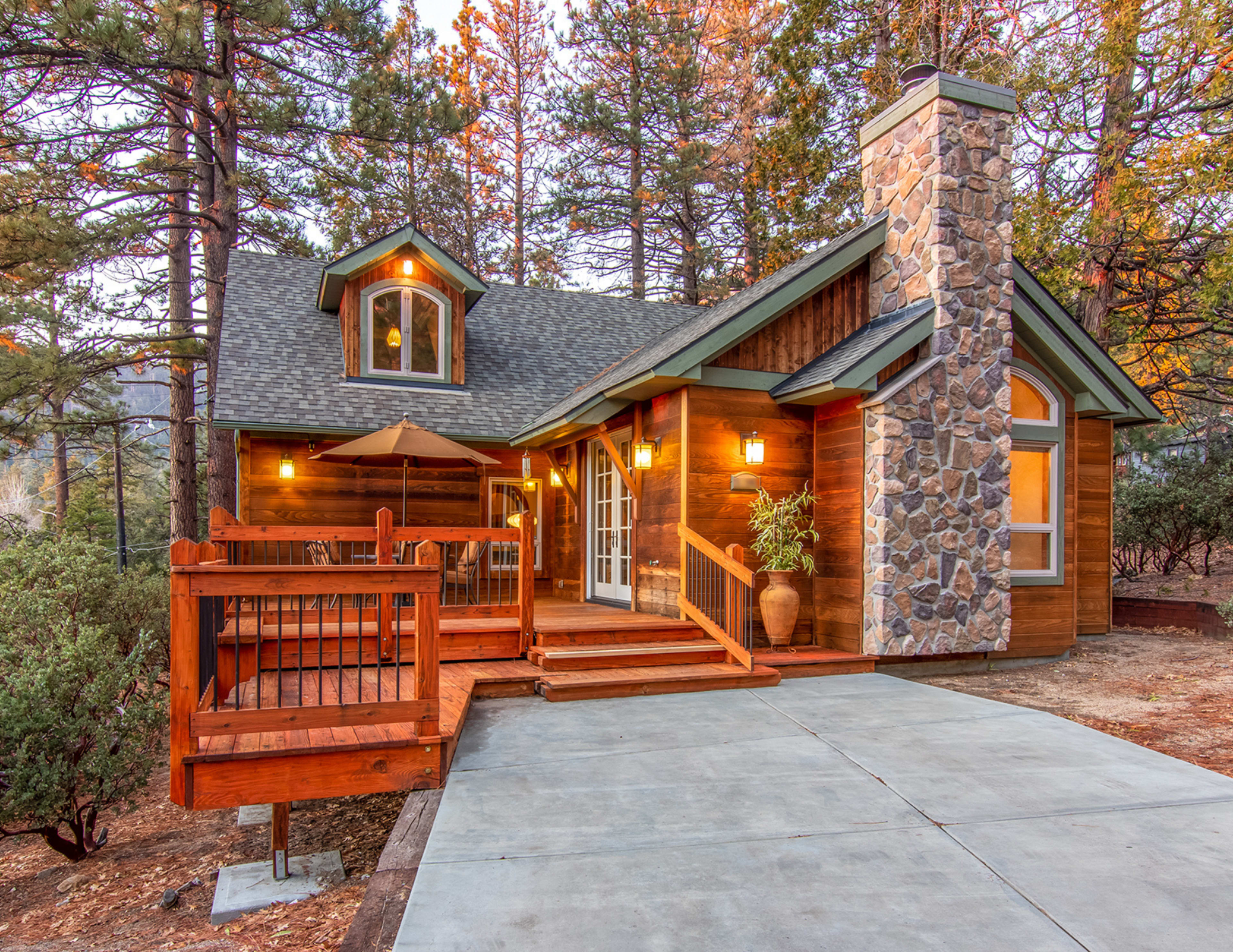 A wooden cabin with a stone chimney features a deck and a concrete driveway, surrounded by trees.