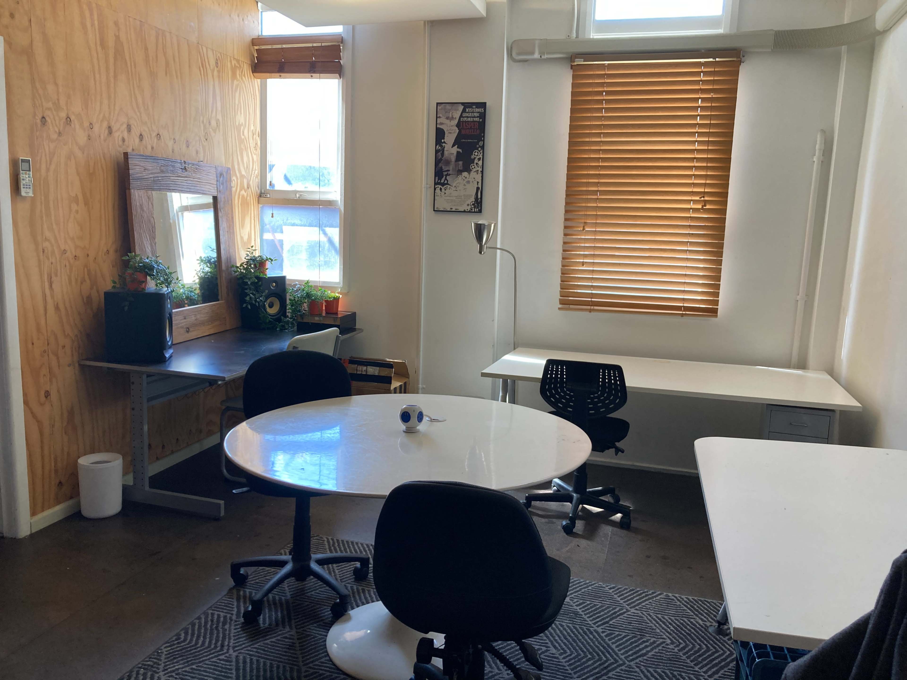The image shows an office space with a wooden wall, two desks, a round table, plants on a shelf, and window blinds.