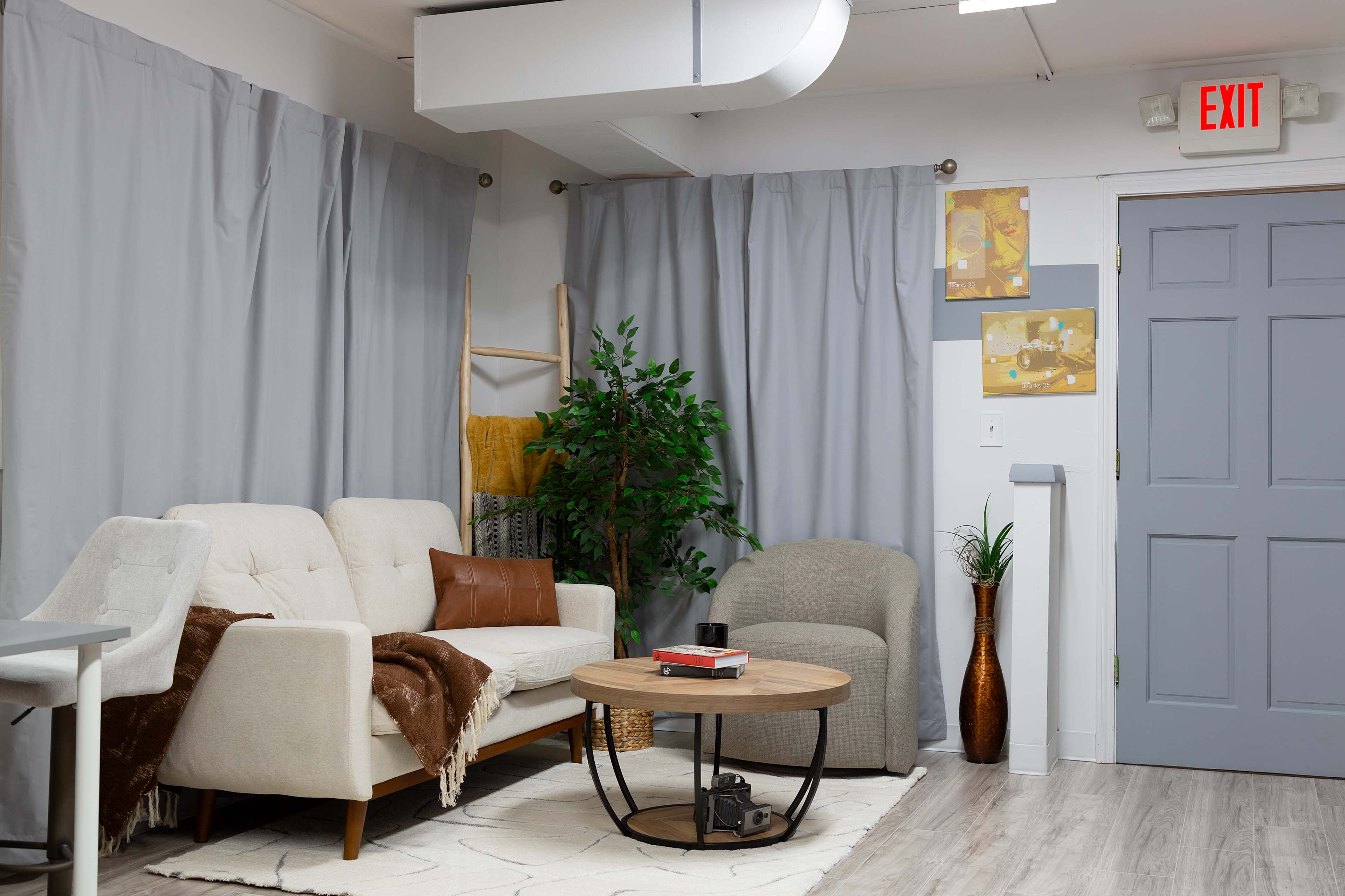 The image shows a living space featuring a light-colored sofa, a round wooden coffee table, a gray armchair, and a plant, with gray curtains covering the windows and art on the walls.
