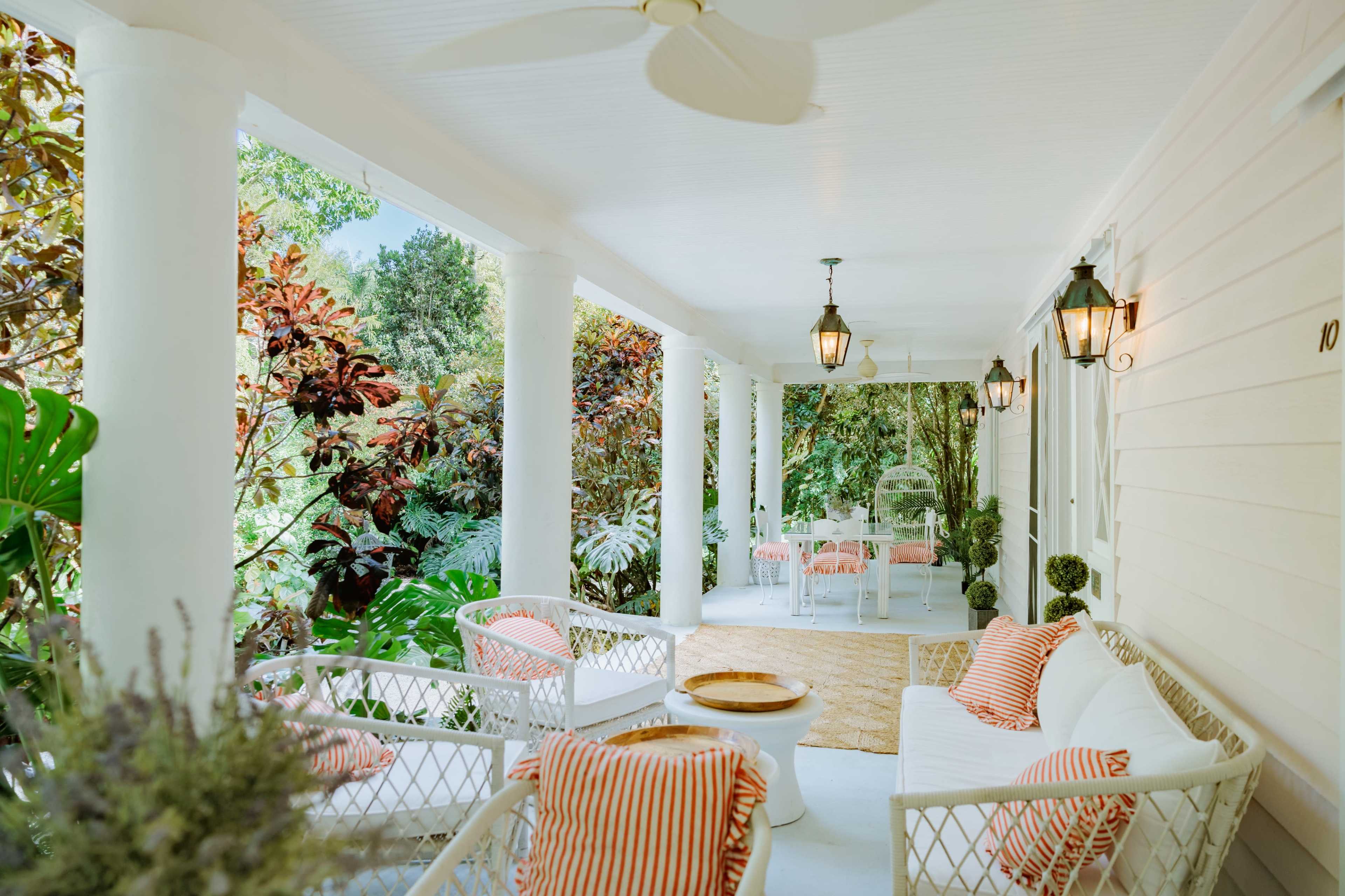 The image shows a covered porch with white wicker furniture and orange-striped pillows, surrounded by lush greenery and hanging lanterns.
