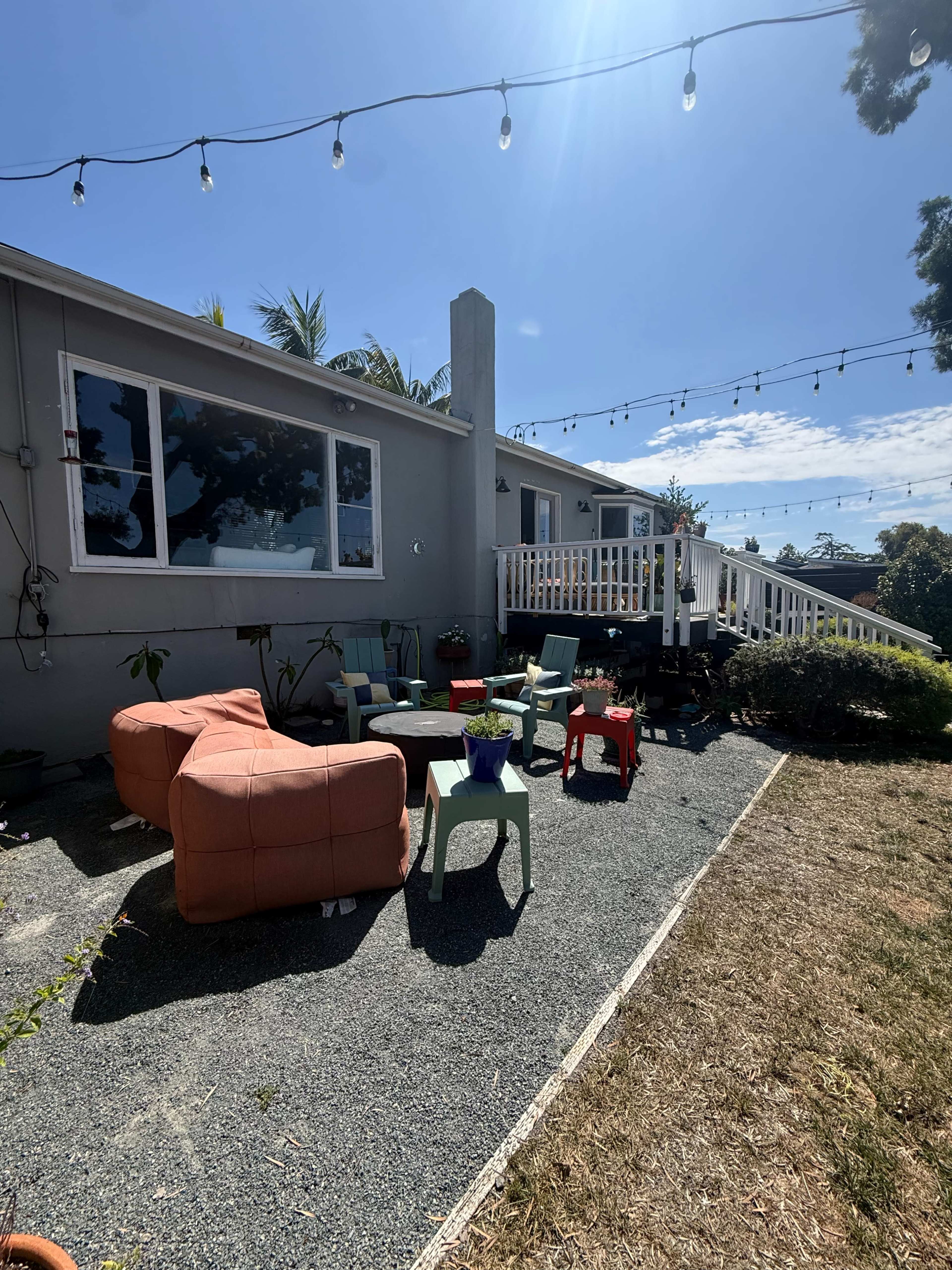 The image shows a backyard with colorful seating arrangements, a gravel path, and string lights overhead, leading to a house with large windows.
