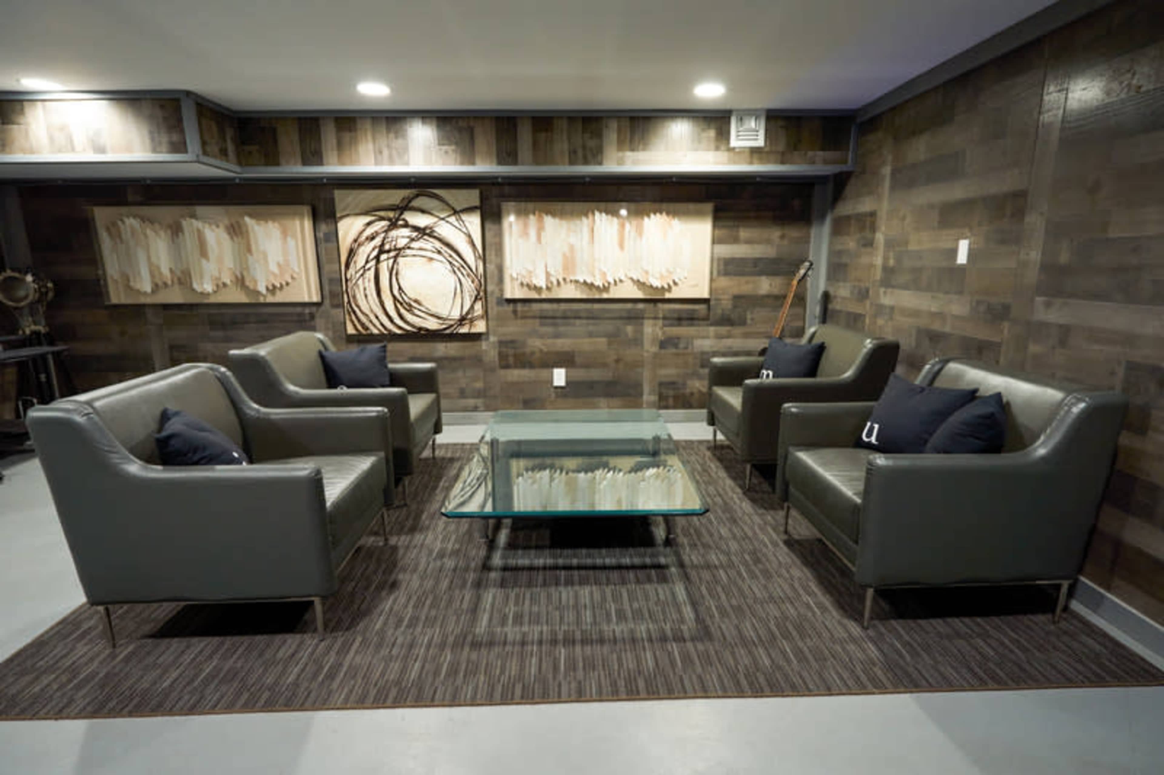 A modern lounge area with four gray leather chairs arranged around a glass-topped coffee table on a patterned rug beneath framed artwork on the wooden walls.