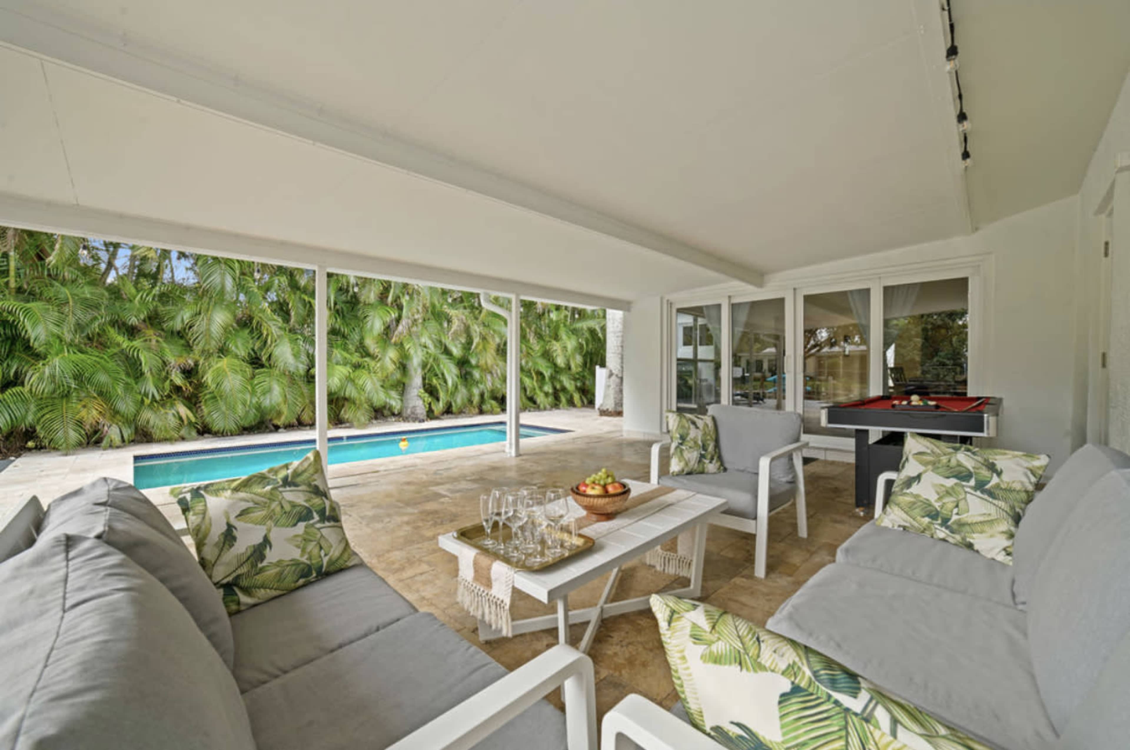 A covered outdoor seating area features gray sofas with floral-patterned cushions and a coffee table set with glasses and fruit, overlooking a swimming pool surrounded by lush greenery.