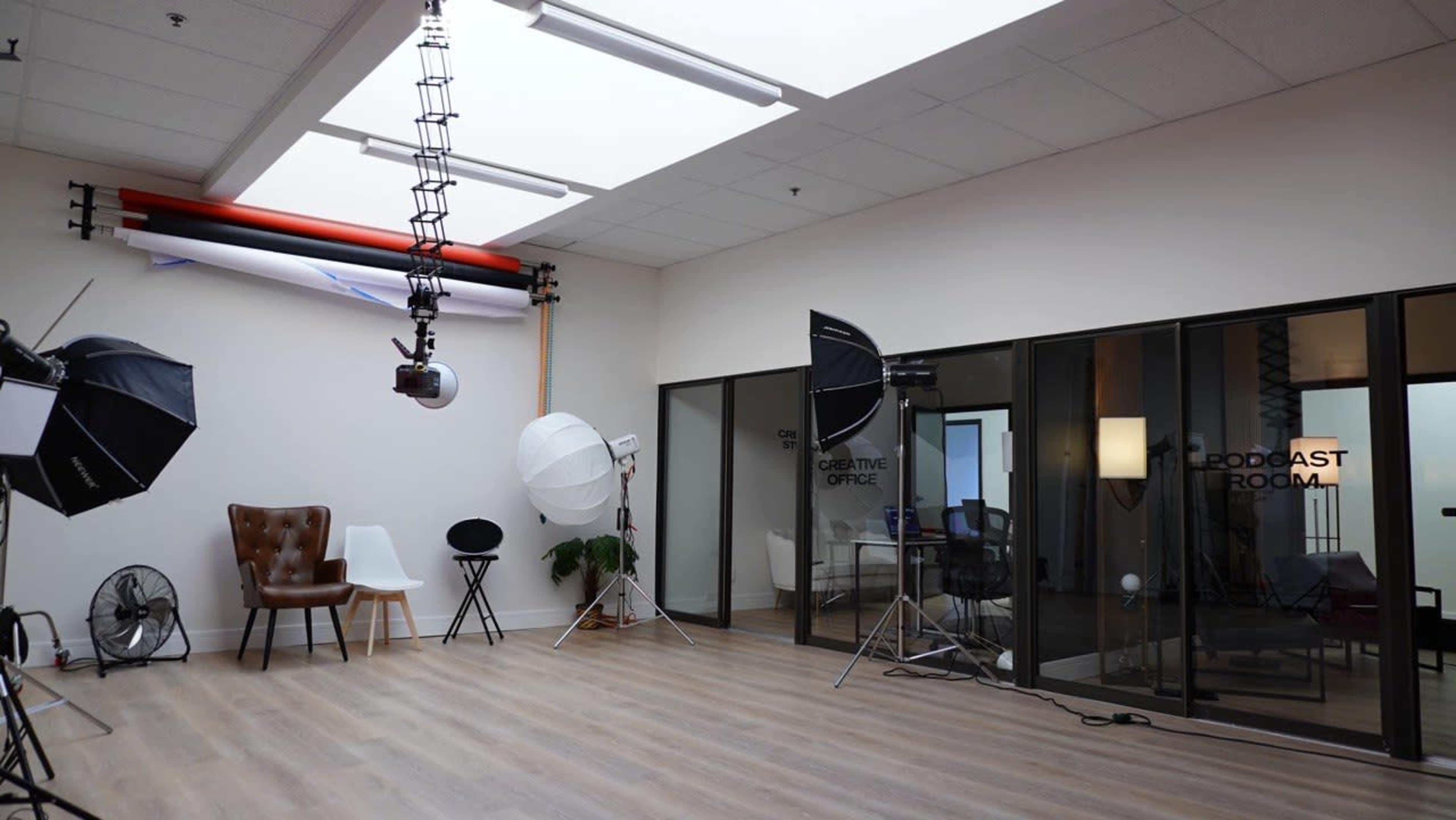 The image shows a spacious studio room with a light wooden floor, featuring a brown leather chair, a white table, photography lighting equipment, and glass doors labeled "PODCAST ROOM."