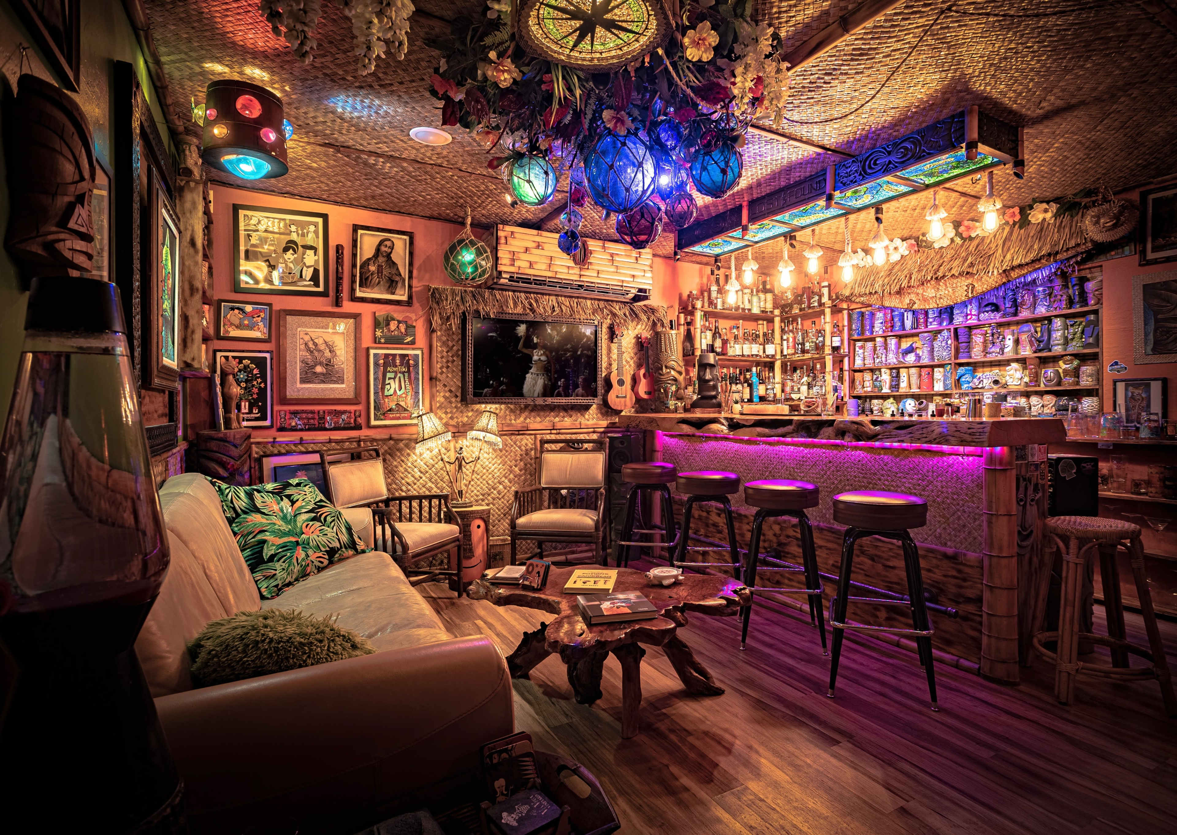 The image depicts a cozy, tropical-themed bar area with a seating arrangement, illuminated by colorful lights and adorned with tiki decor.