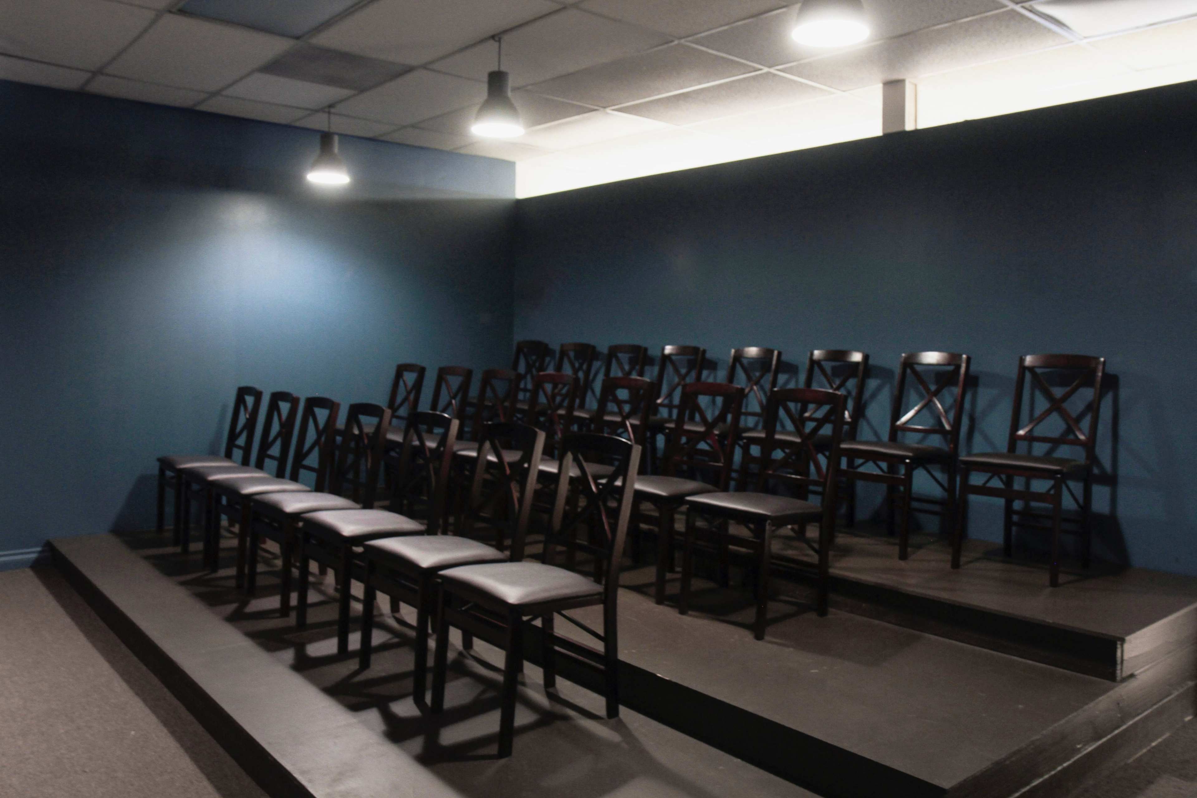 A set of black wooden chairs is arranged in rows on a raised platform against a blue wall under overhead lights.