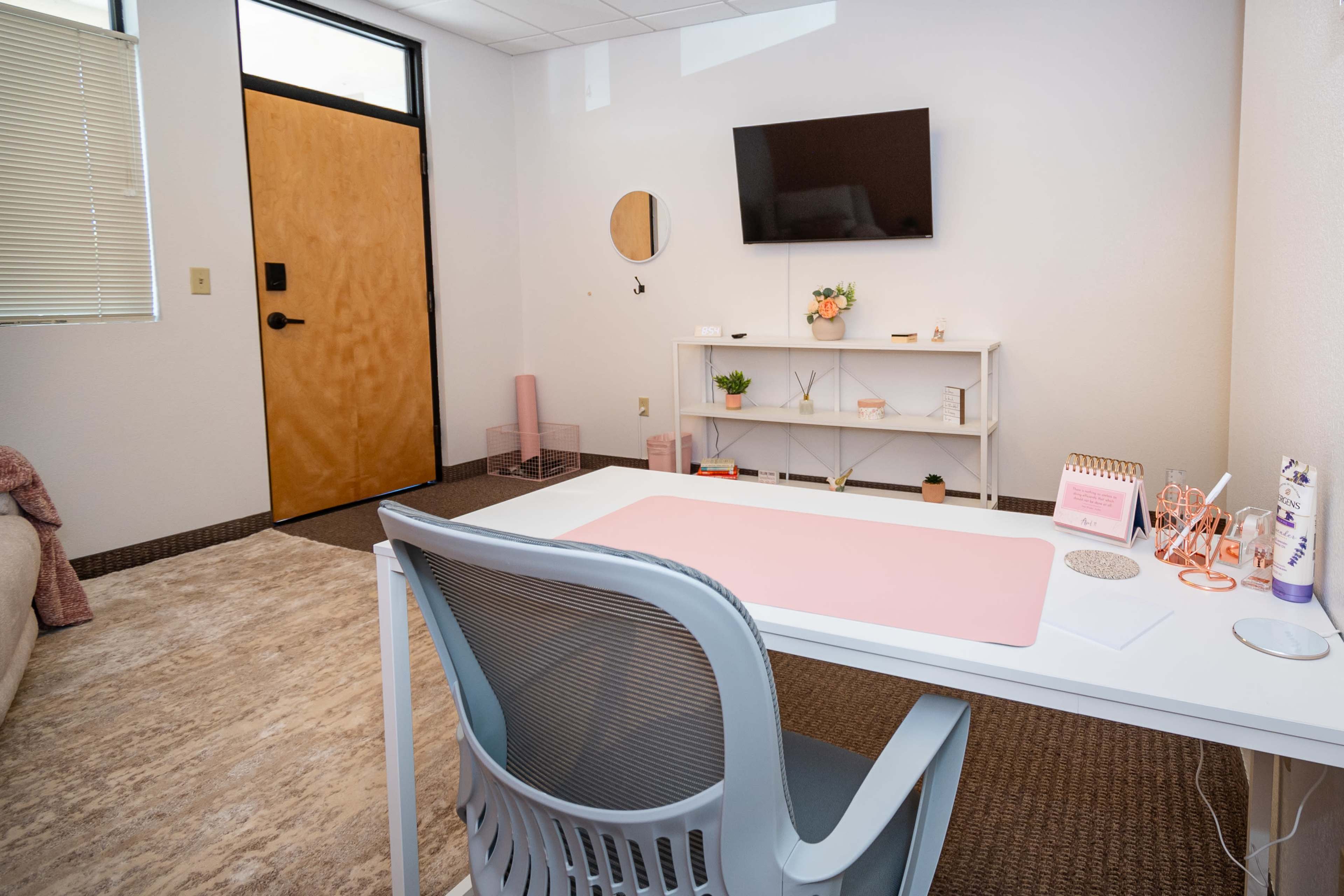The image shows a small office space featuring a white desk with a pink mat, a television mounted on the wall, and a shelving unit with decorative items.