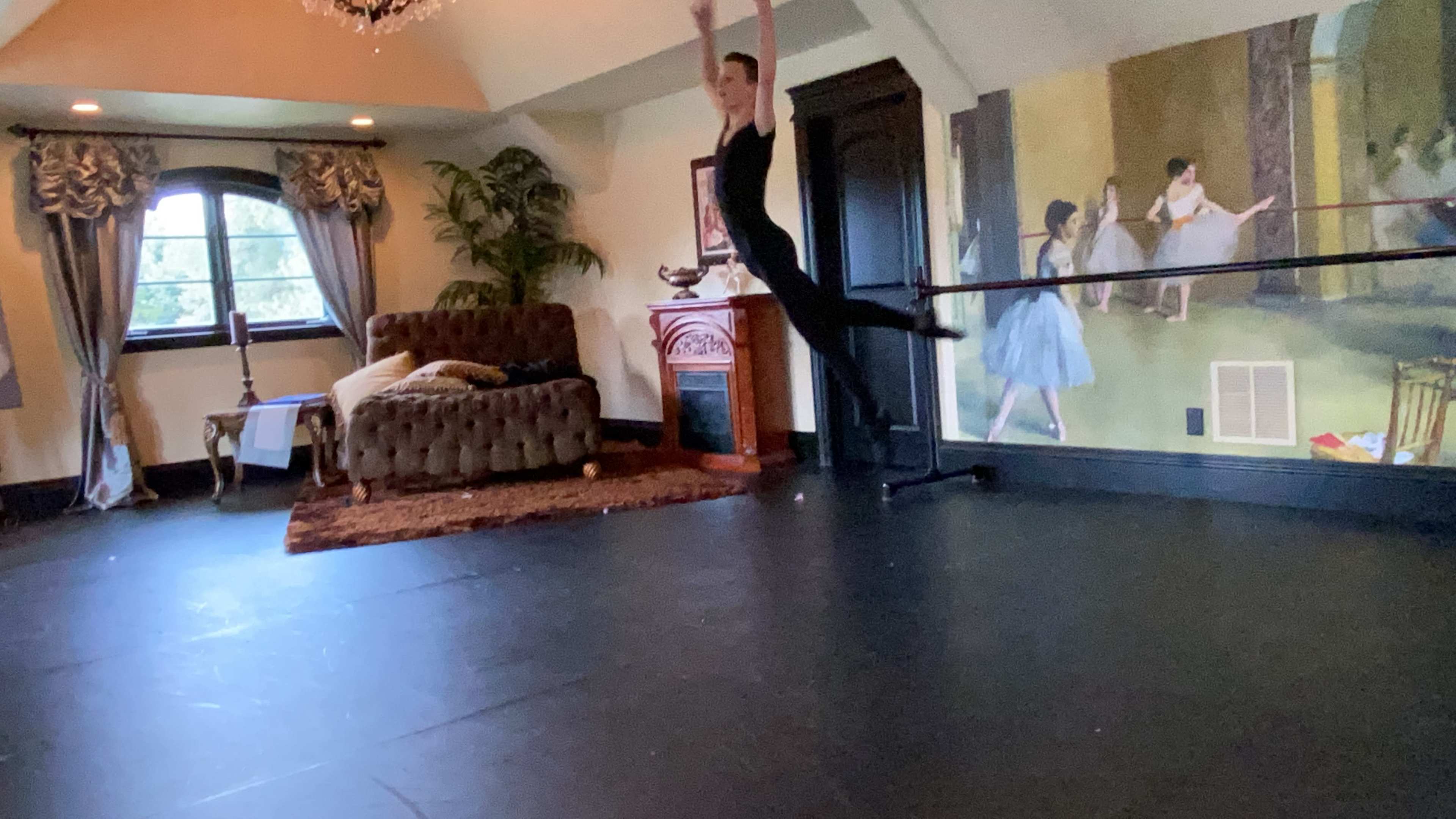 A dancer performs a leap in a studio decorated with elegant furnishings and a mural depicting ballet scenes.