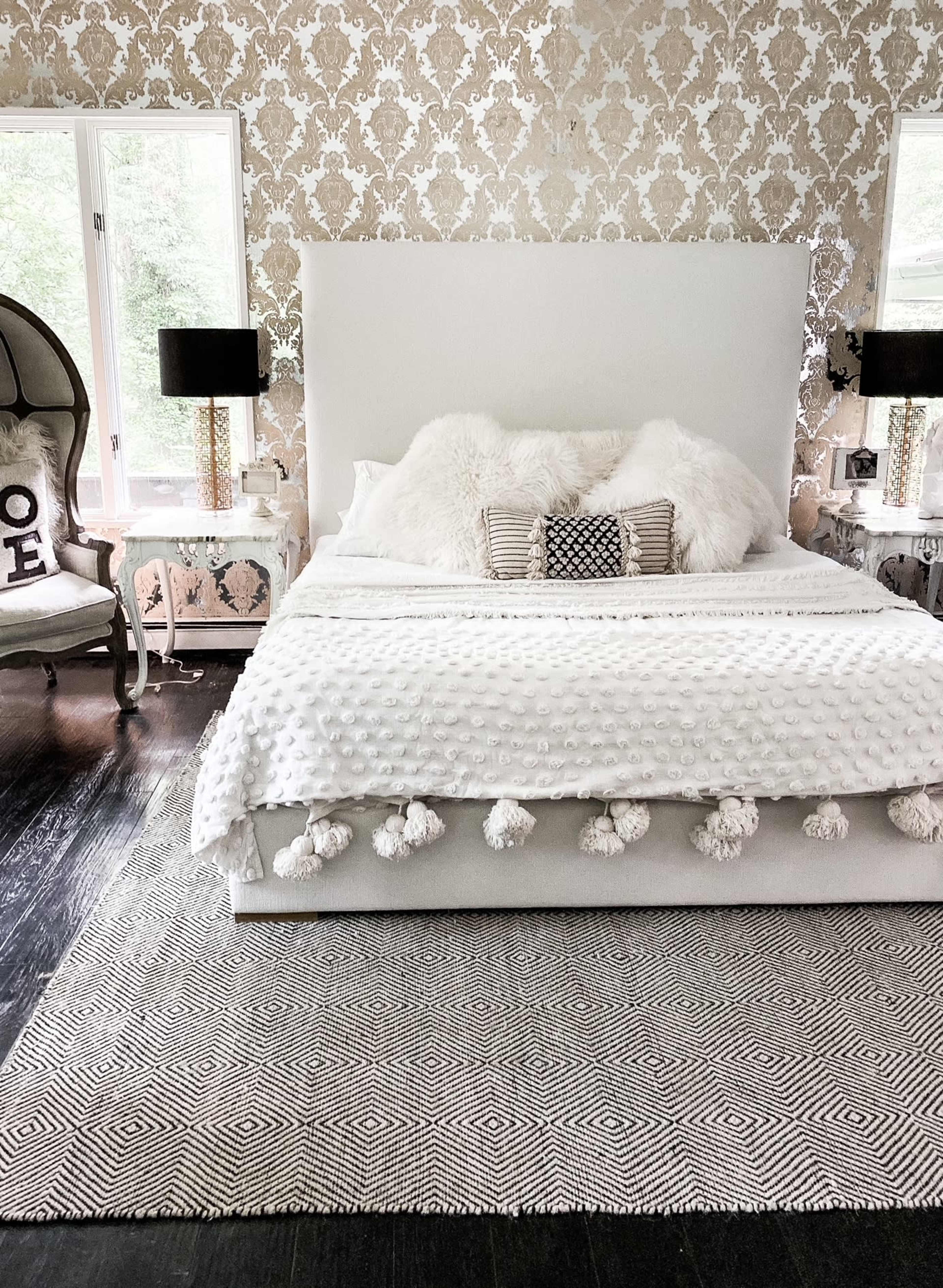 A neatly arranged bedroom with a large bed covered in a textured white blanket and decorative pillows, flanked by two lamps on nightstands, against a patterned wallpaper background.