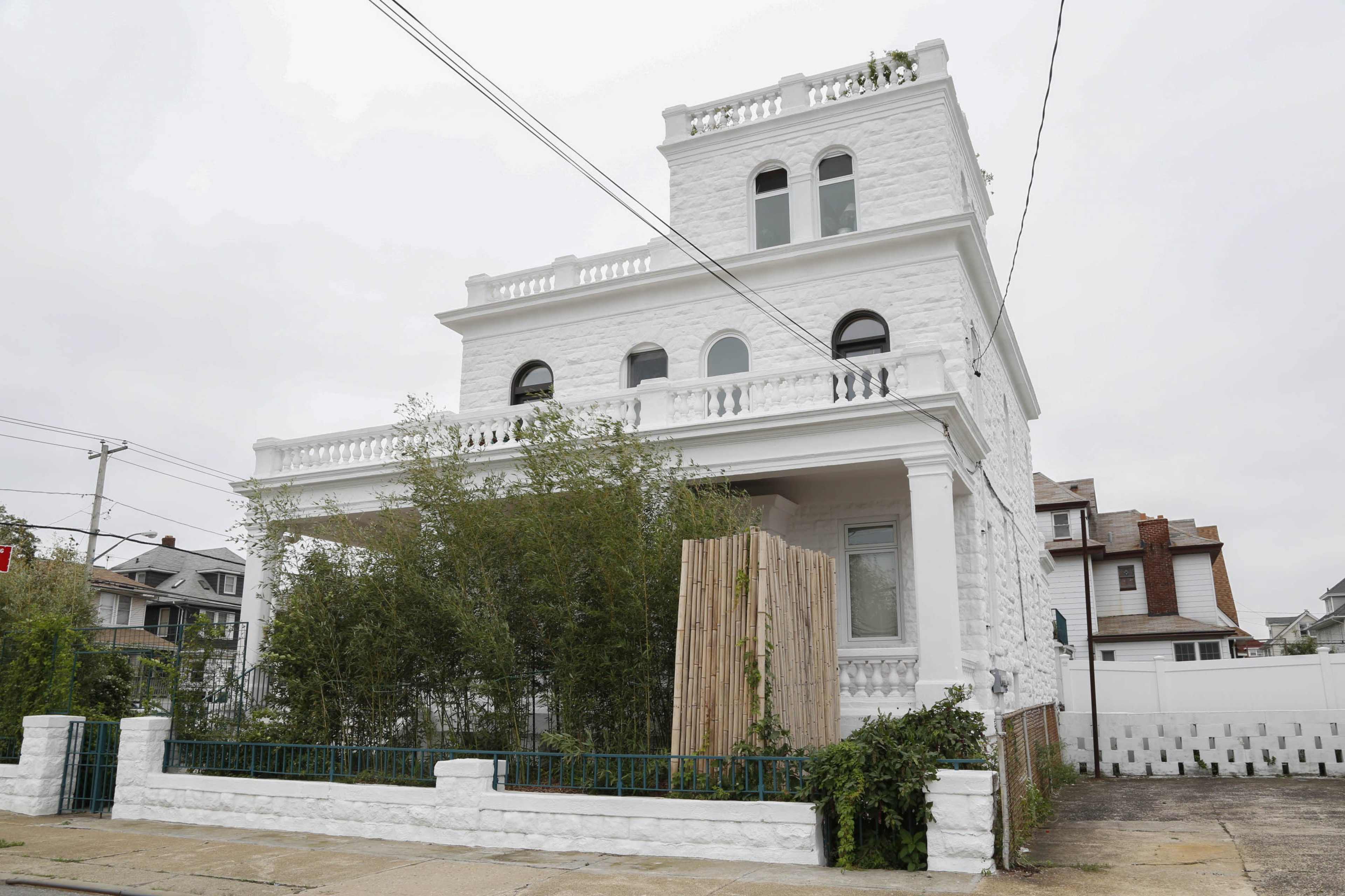 A large, white, three-story house features a balcony and a bamboo fence in the front yard.