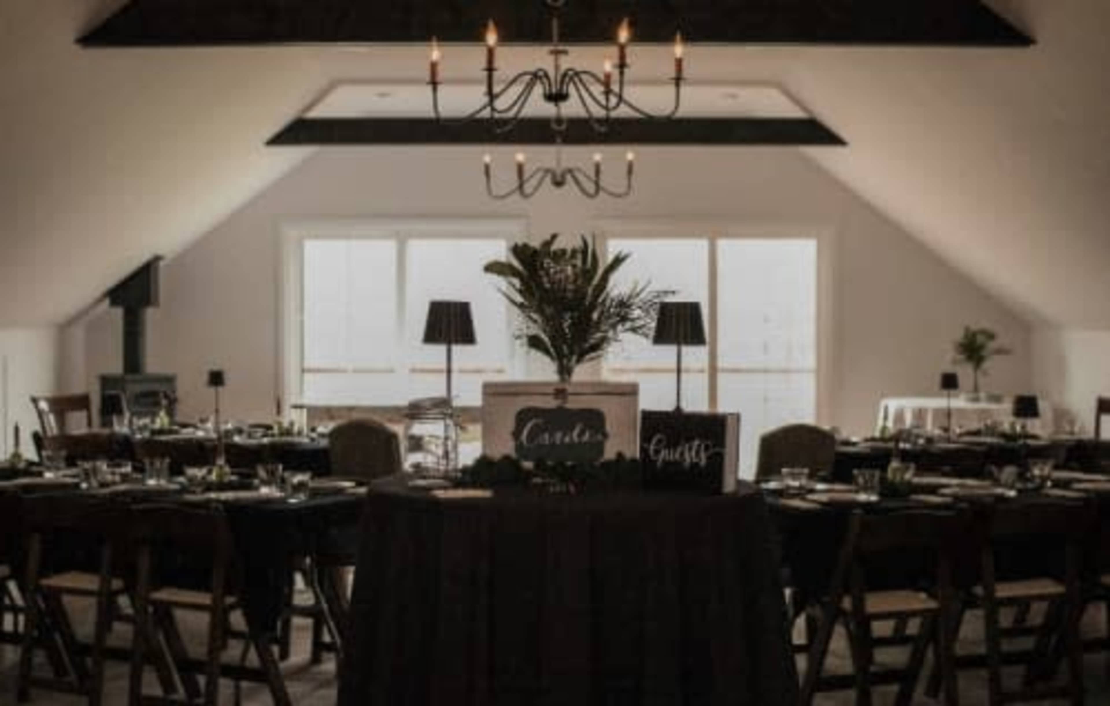 A dining area features neatly arranged tables with dark tablecloths, chairs, and a centerpiece under a chandelier.