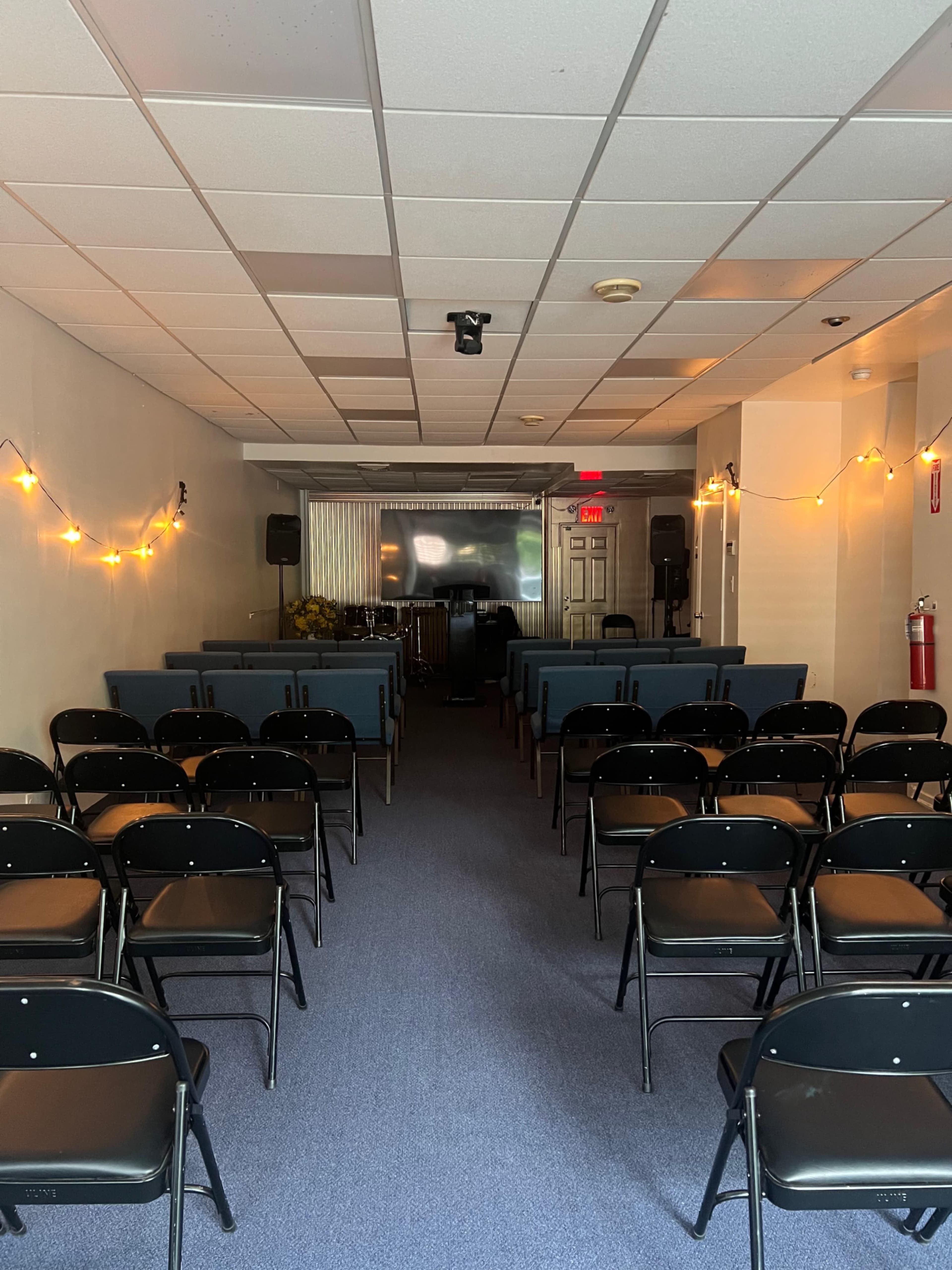 The image shows a linear arrangement of black chairs in a small, well-lit room with a projector screen at the front and soft lighting.