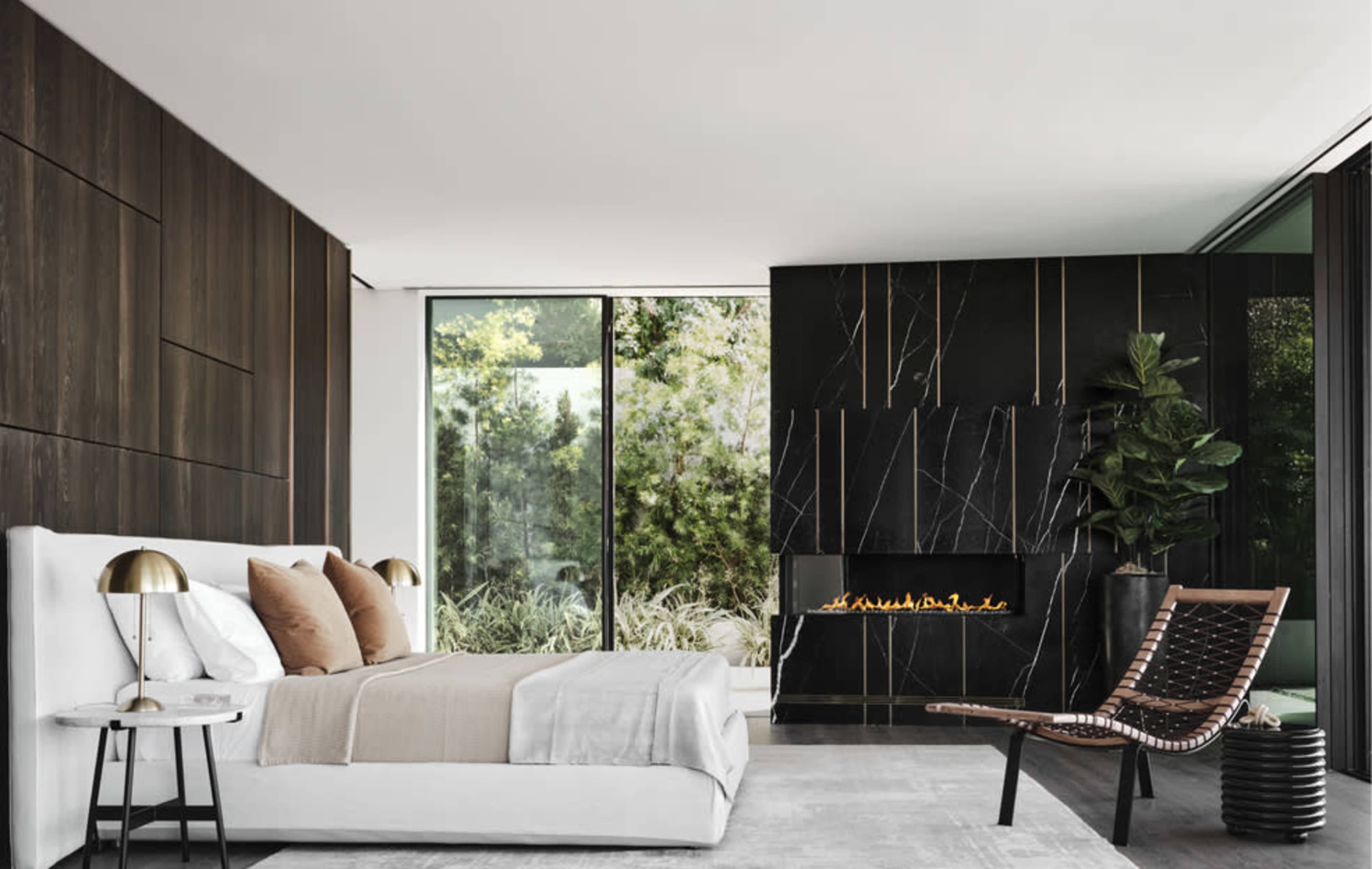 A modern bedroom featuring a large bed with decorative pillows, a minimalist side table, a sleek fireplace, and large windows facing a green outdoor view.