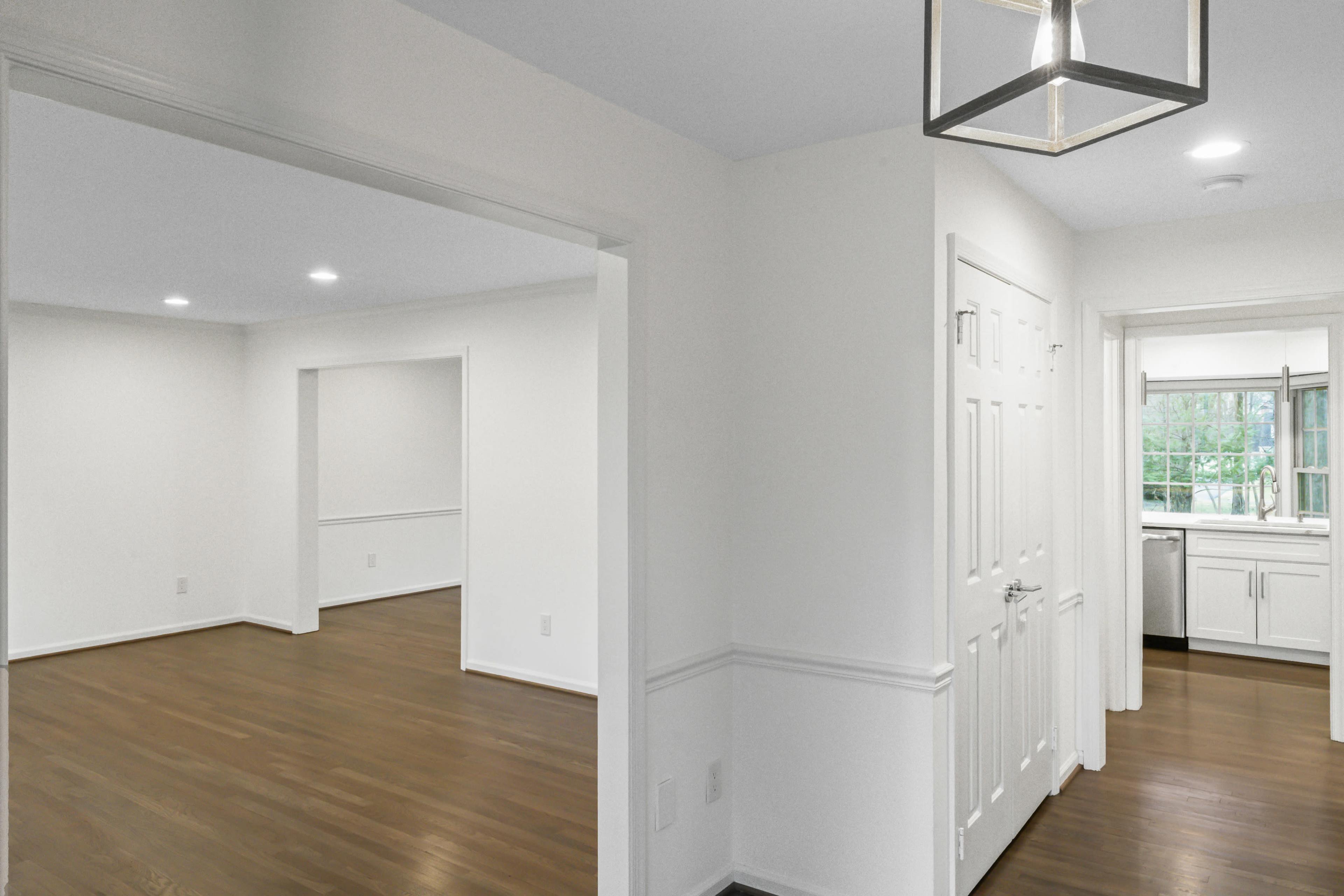 The image shows a well-lit hallway leading to a kitchen, with a view of adjacent rooms featuring hardwood floors and white walls.