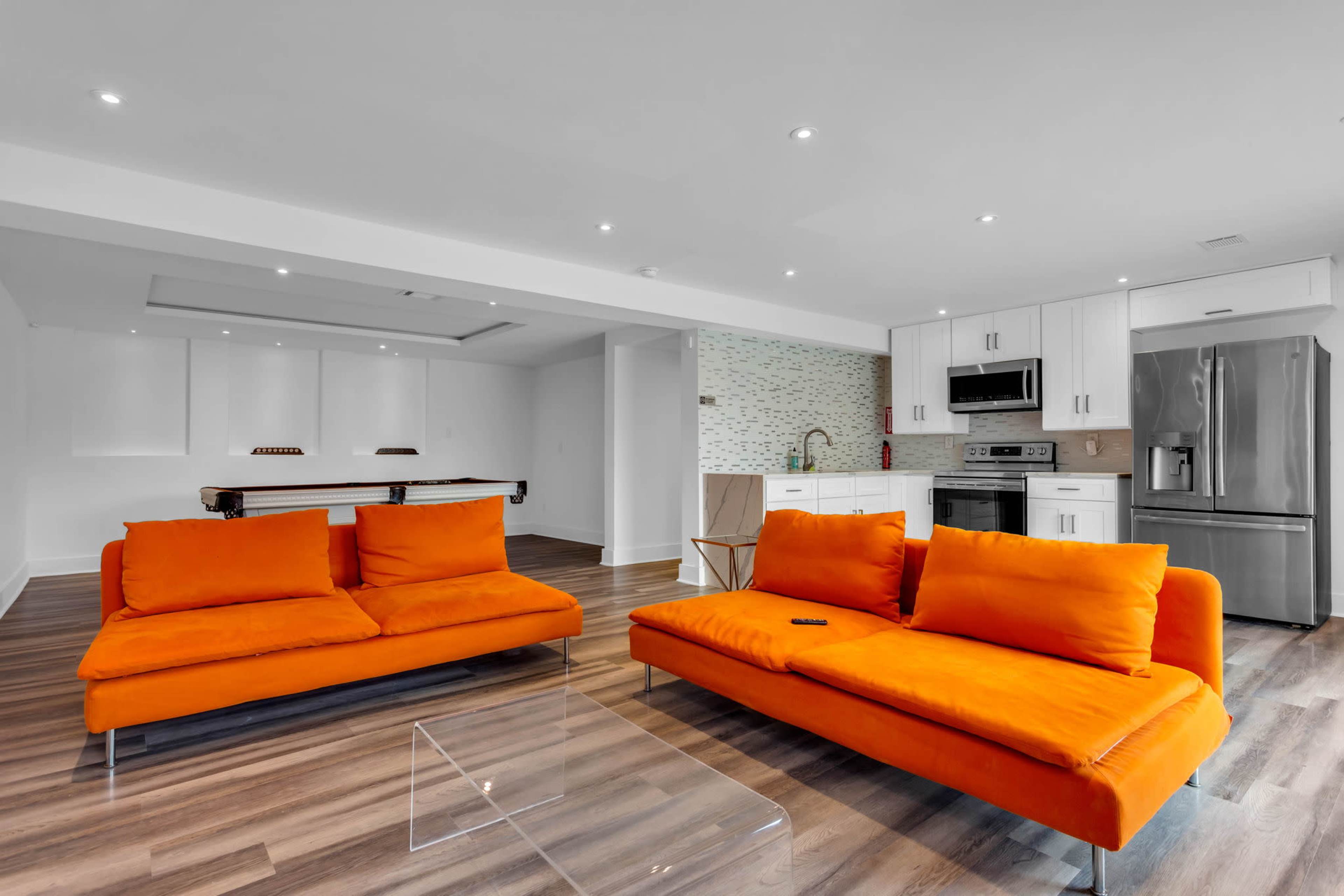 A modern living space features two orange sofas facing a kitchen with stainless steel appliances and a pool table in the background.