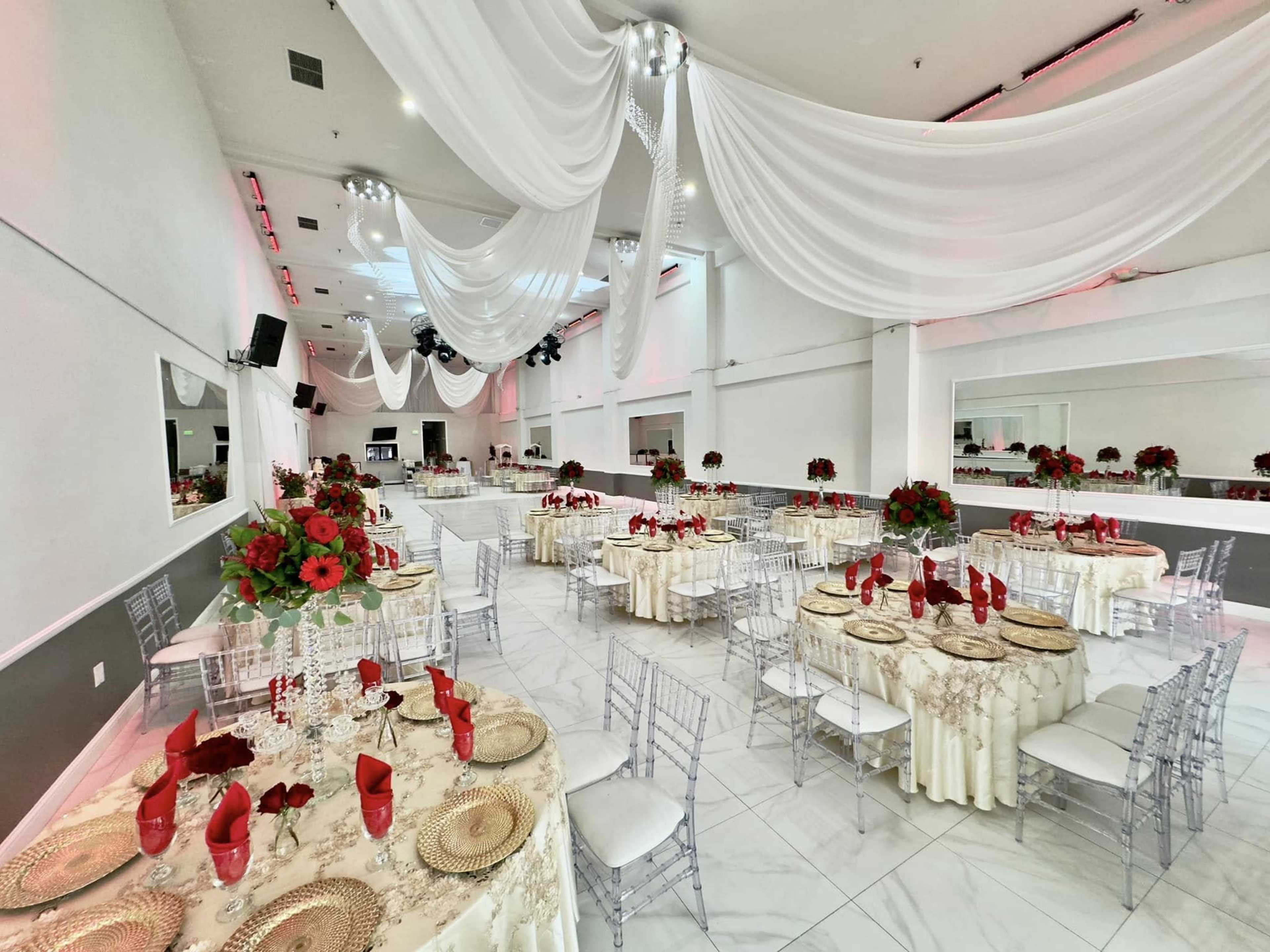 The image shows a spacious banquet hall with elegantly arranged tables, each adorned with red floral centerpieces and white tablecloths under draped white fabric and soft lighting.