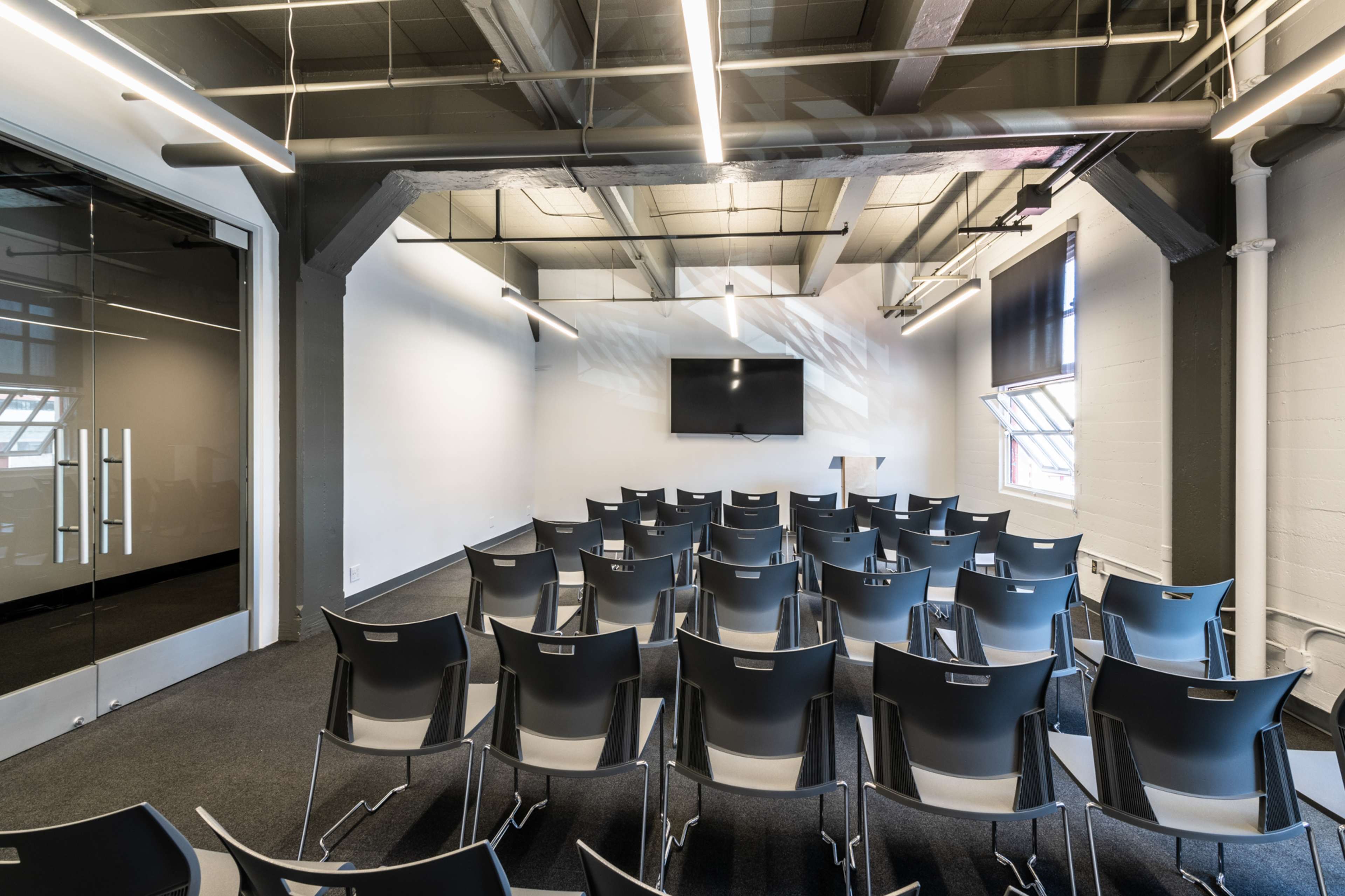 An empty conference room with rows of black chairs facing a wall-mounted screen, lit by overhead fluorescent lights.
