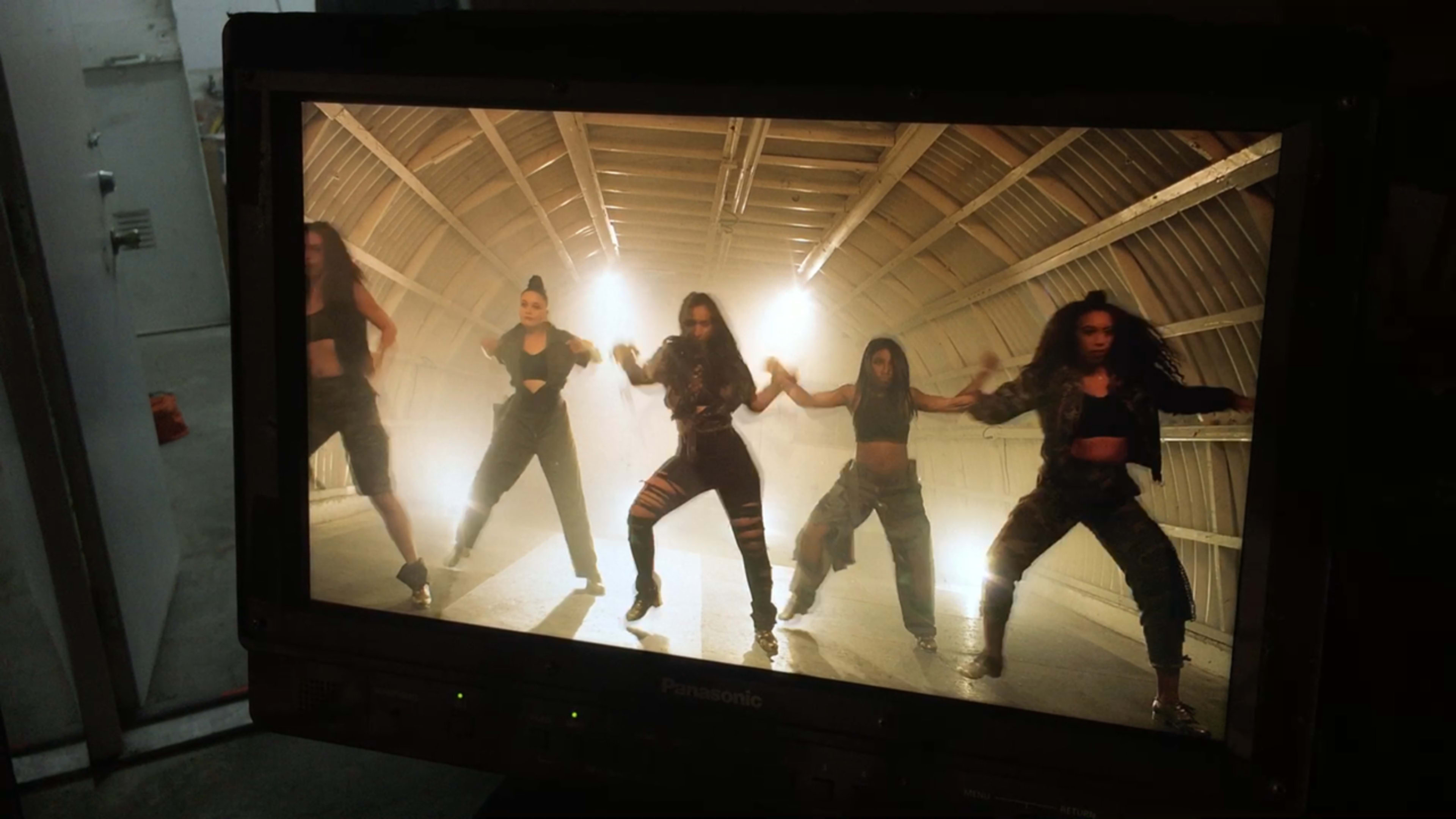 A group of five dancers performs synchronized movements in a dimly lit studio with a wooden ceiling.