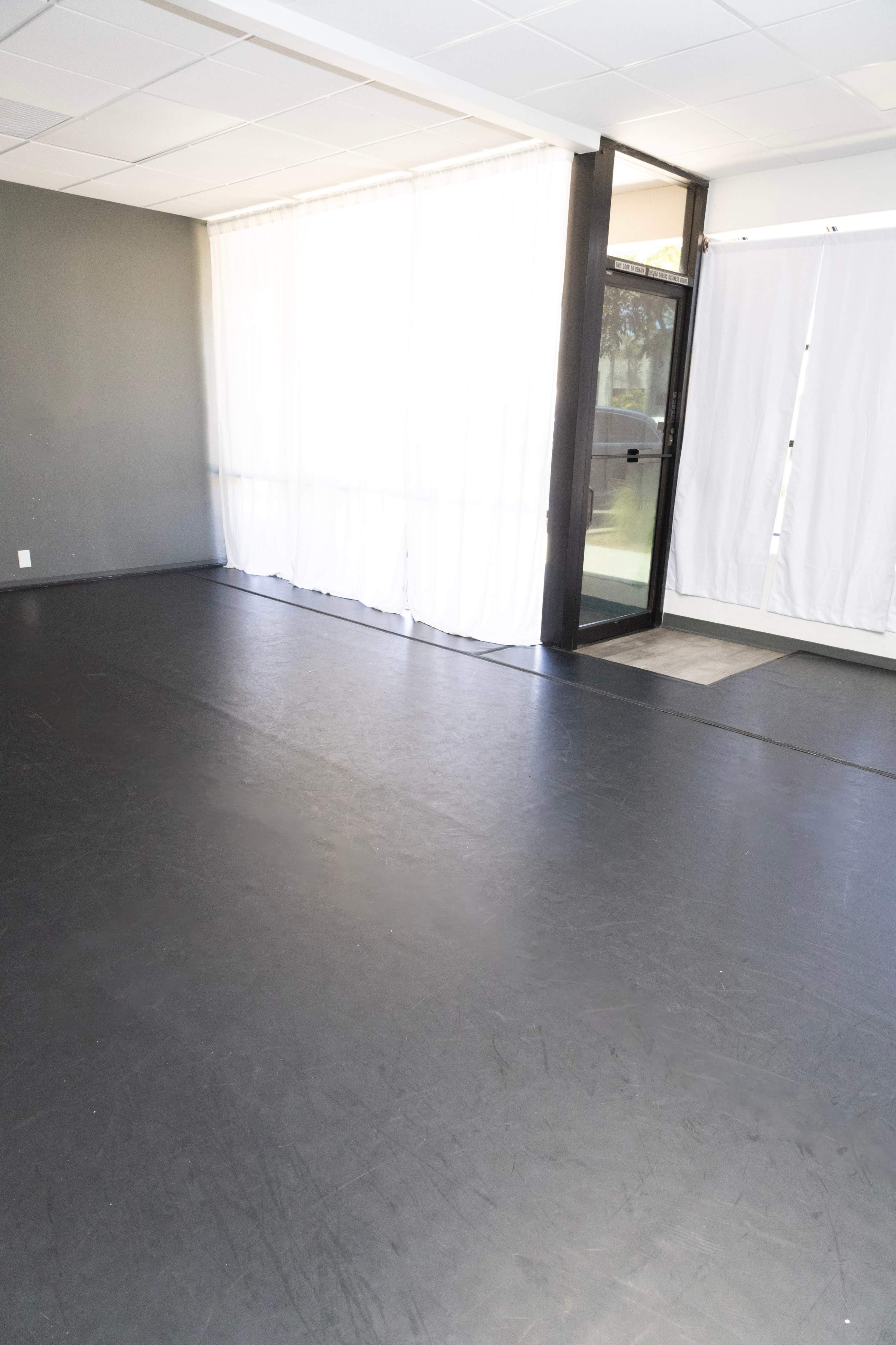 The image shows an empty room with a dark floor, large windows covered by sheer curtains, and a door leading outside.