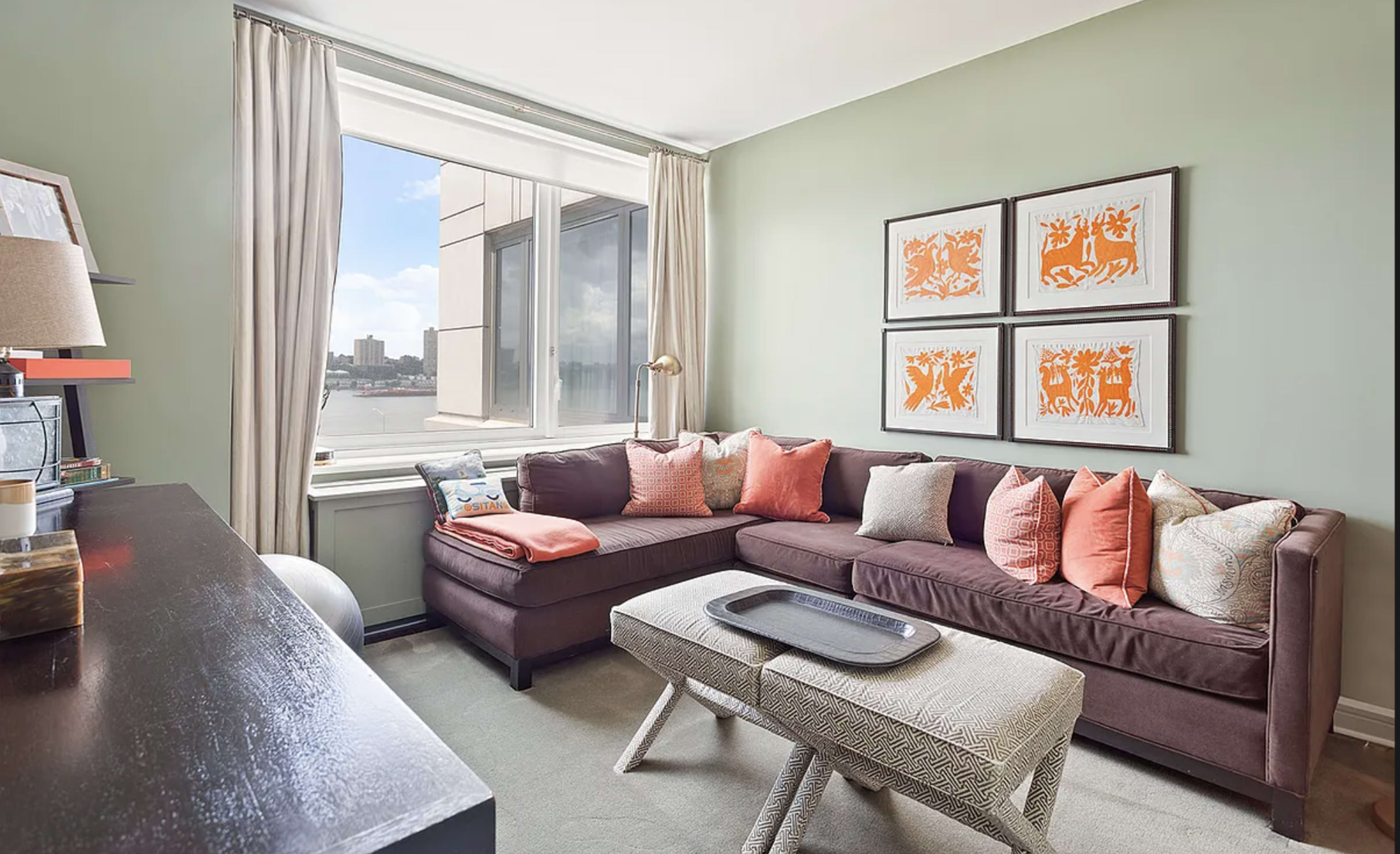 A small living room with a brown sectional sofa adorned with decorative pillows, a light-colored coffee table, and framed art on the walls, all set against a green backdrop and a large window.