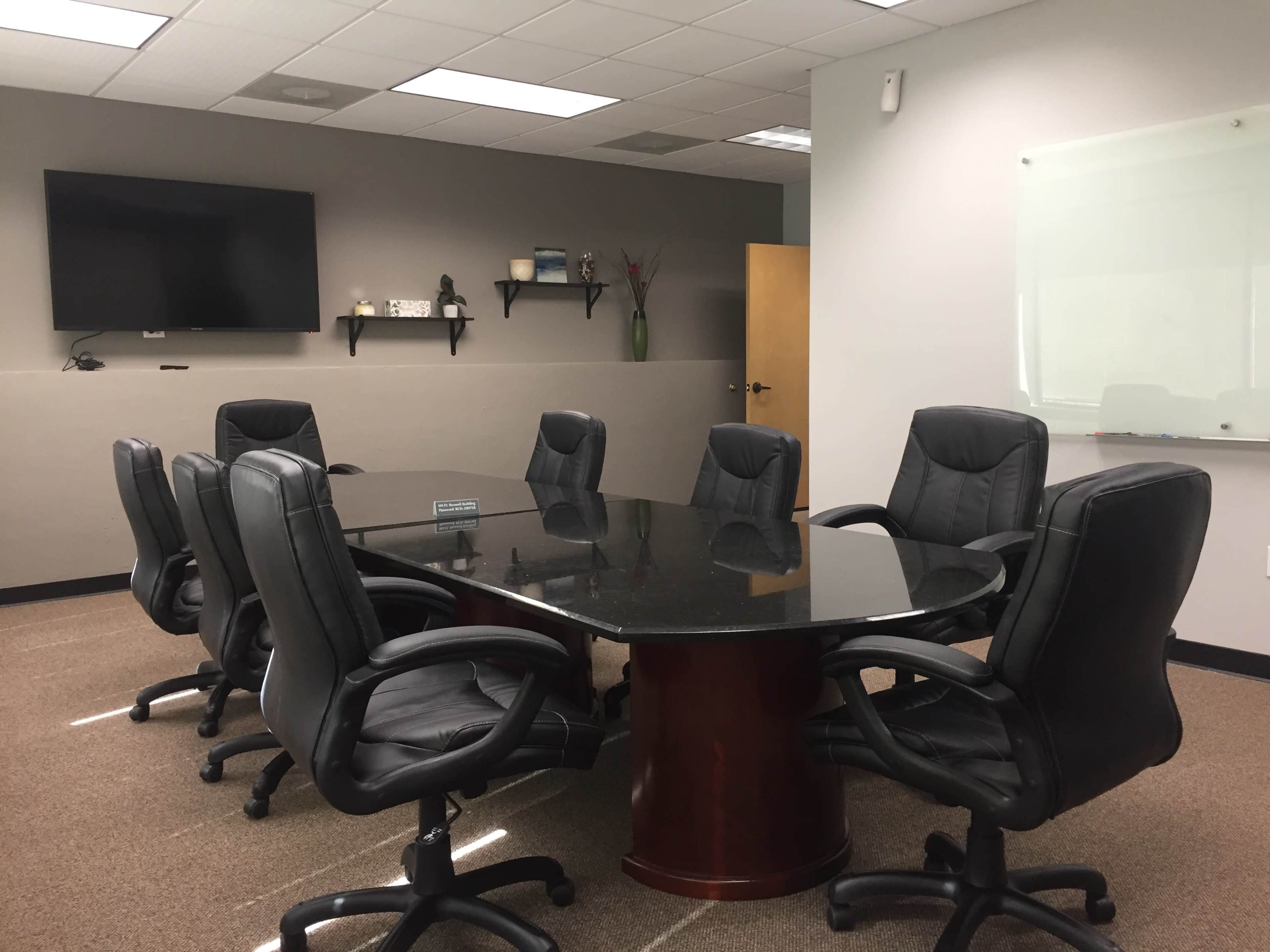 The image shows a conference room with a large round table surrounded by black rolling chairs and a wall-mounted television.
