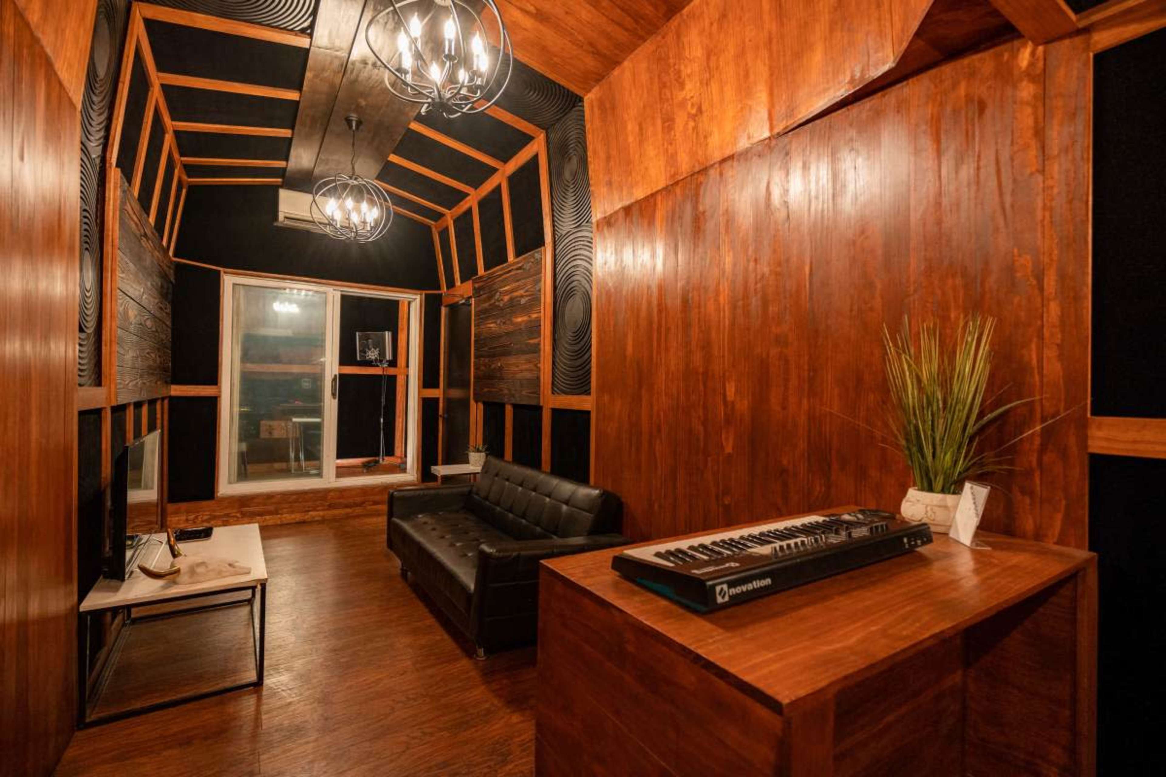 The image depicts a wooden-paneled music studio featuring a black sofa, a keyboard on a table, and large windows providing natural light.